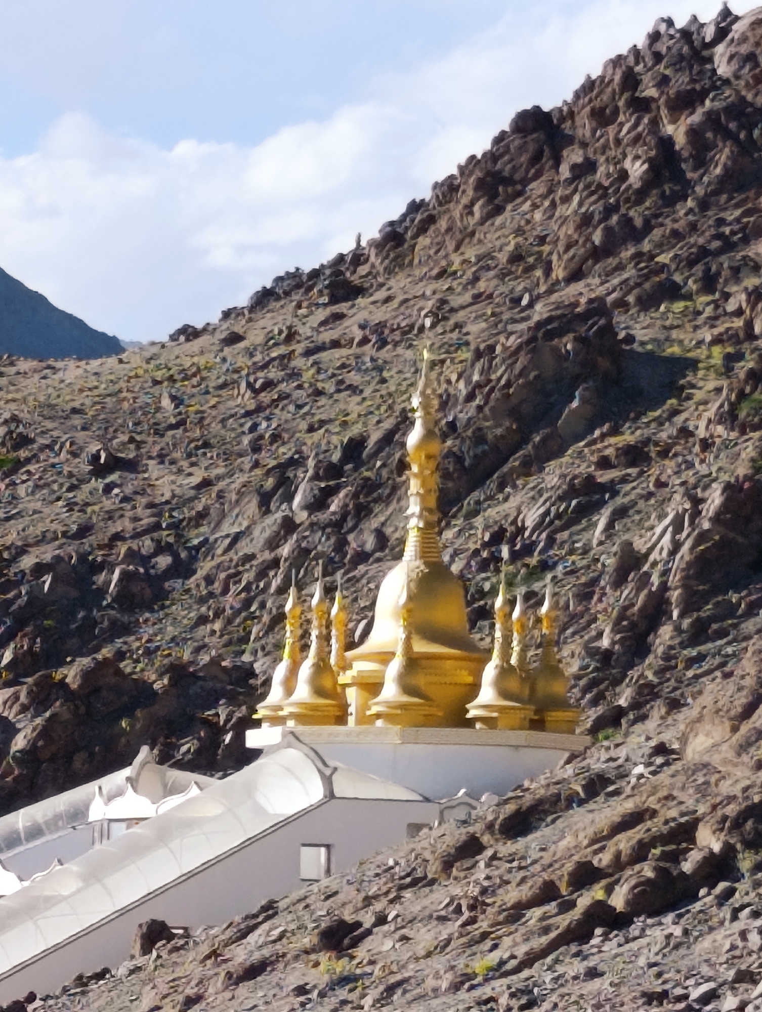 The image shows a Buddhist stupa complex nestled against a rocky mountainside in Shey, India. The stupa, adorned with golden spires, stands in stark contrast to the rugged, barren terrain. A translucent white roof structure extends from the base of the stupa, partially covering the ground in front of it. The sky above is a pale blue with scattered white clouds. The rocky slopes are covered in loose stones and sparse vegetation, indicative of a high-altitude environment. The overall impression is one of spiritual serenity amidst a harsh, natural landscape.