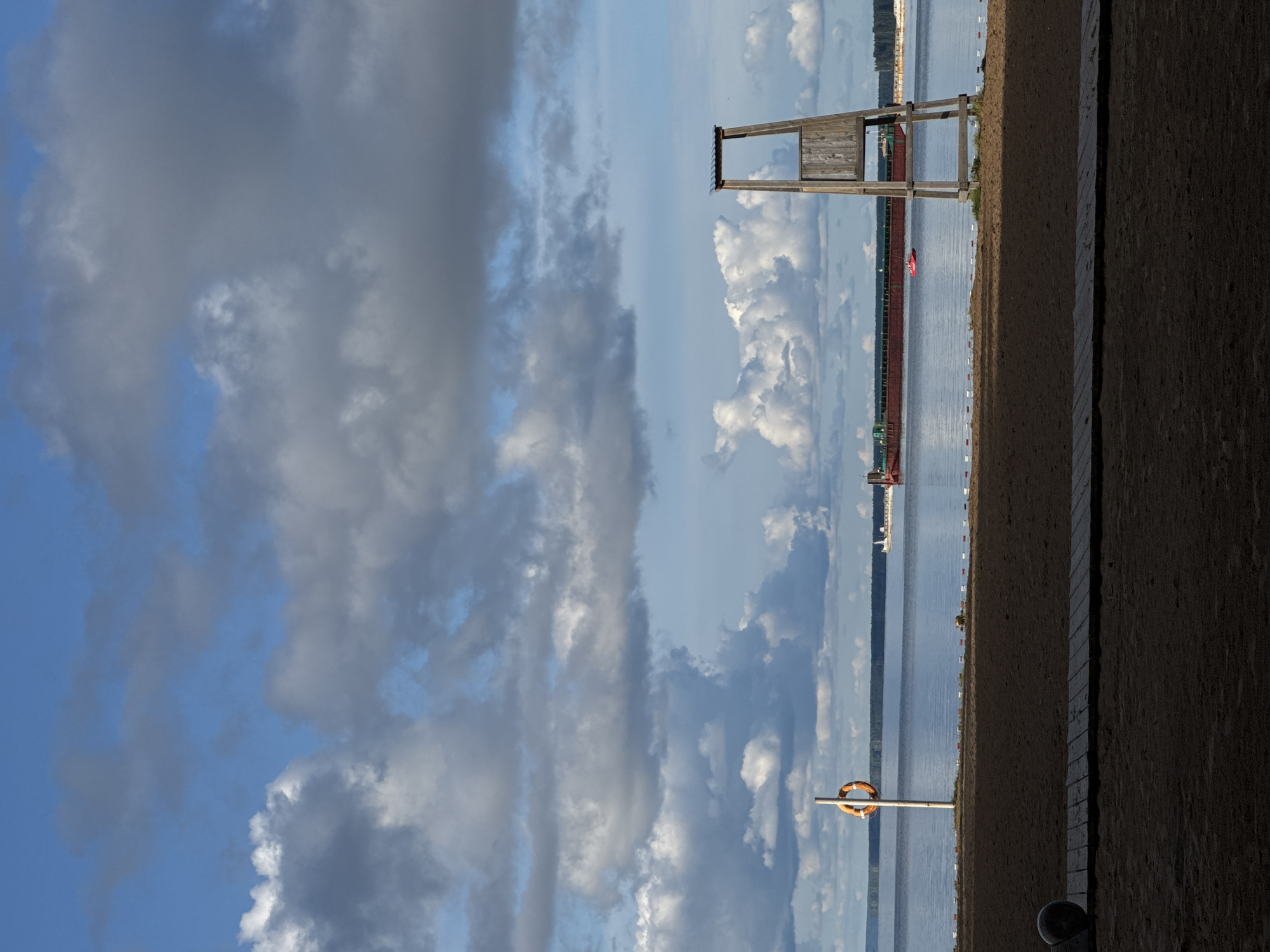 The image depicts a serene daytime scene at a beach in Chaikovsky, Russia, overlooking the Kama River. In the foreground, a sandy beach is visible with a wooden walkway stretching across it. A black, round object, possibly a ball, rests on the sand to the left. A lifebuoy hangs on a pole to the left of the frame, near the water's edge.
In the midground, the calm, blue water of the Kama River stretches towards the horizon. A large, red cargo ship is docked or moored on the water, appearing distant. A small, bright red boat is also visible on the water, closer to the shore than the cargo ship. A wooden lifeguard tower stands prominently on the right side of the beach, overlooking the water. The background features a distant treeline, suggesting a forested area along the riverbank.
The sky above is a dramatic canvas of blue with numerous white and grey clouds, some appearing fluffy and voluminous, others more wispy, creating a sense of dynamic weather. The overall atmosphere is peaceful and invites contemplation of a quiet day by the water.