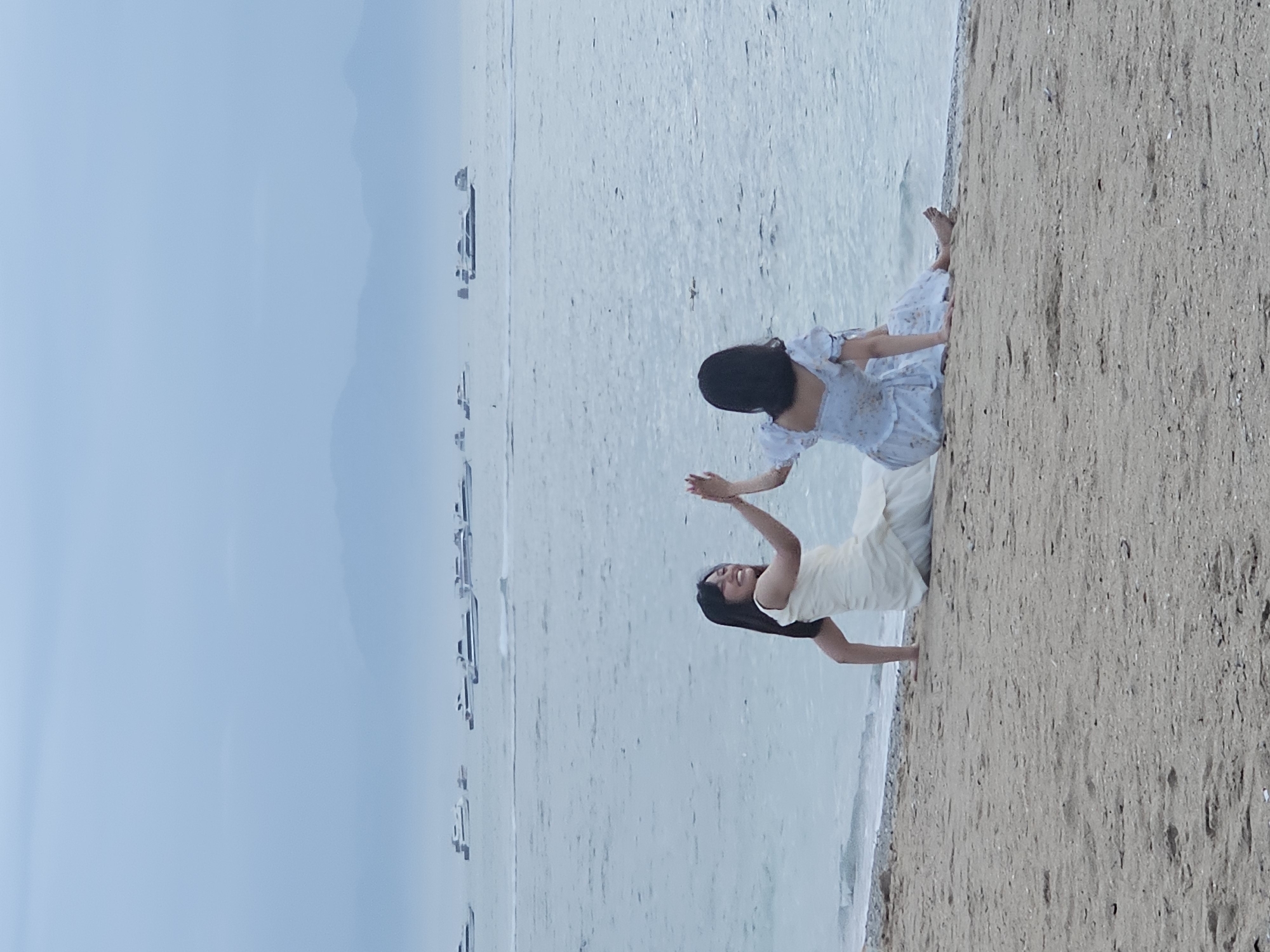Two women are interacting on a beach in Denpasar, Indonesia. One woman is standing, wearing a light-colored dress, her arms outstretched towards the other. The other woman is sitting on the sand, wearing a blue and white floral dress. Behind them is the ocean and a cloudy sky. A beach extends to the right of the women. There are objects in the ocean towards the left.