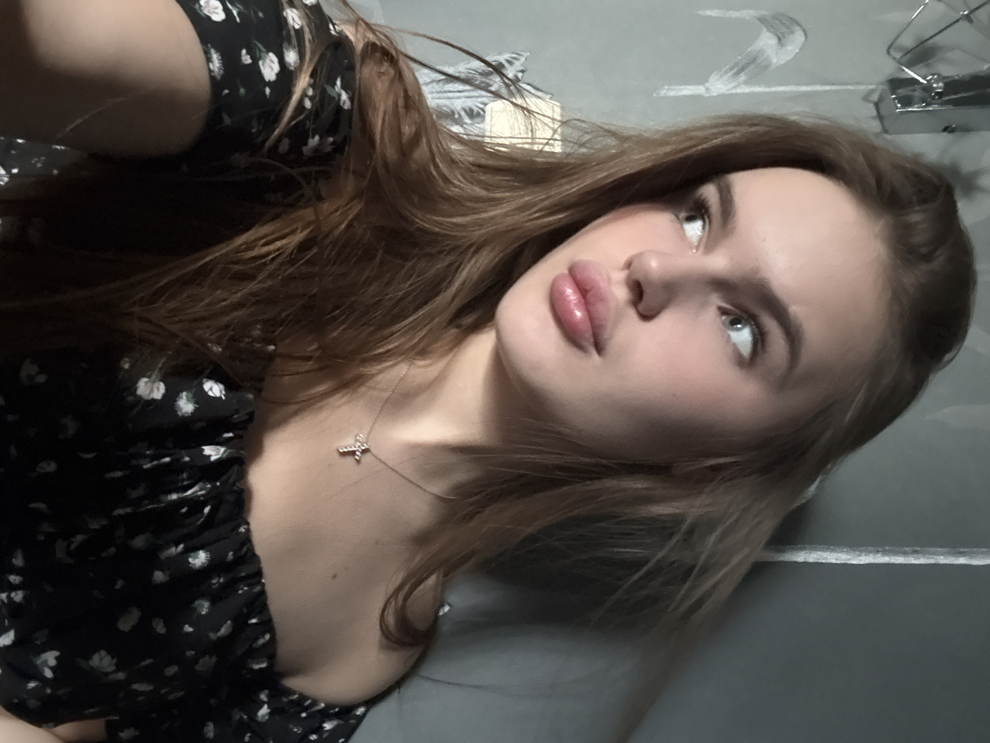 The image shows a young woman with long, brown hair, wearing a black off-the-shoulder top with a floral pattern and a cross necklace. She is looking to her left, with her lips slightly parted. The background appears to be an indoor setting, with a wall that has some light-colored markings on it. A metallic object is visible in the upper left corner of the image. The lighting suggests it might be indoors, possibly in a residential area of Dnipro, Ukraine, given the context provided. The overall mood is casual and relaxed.