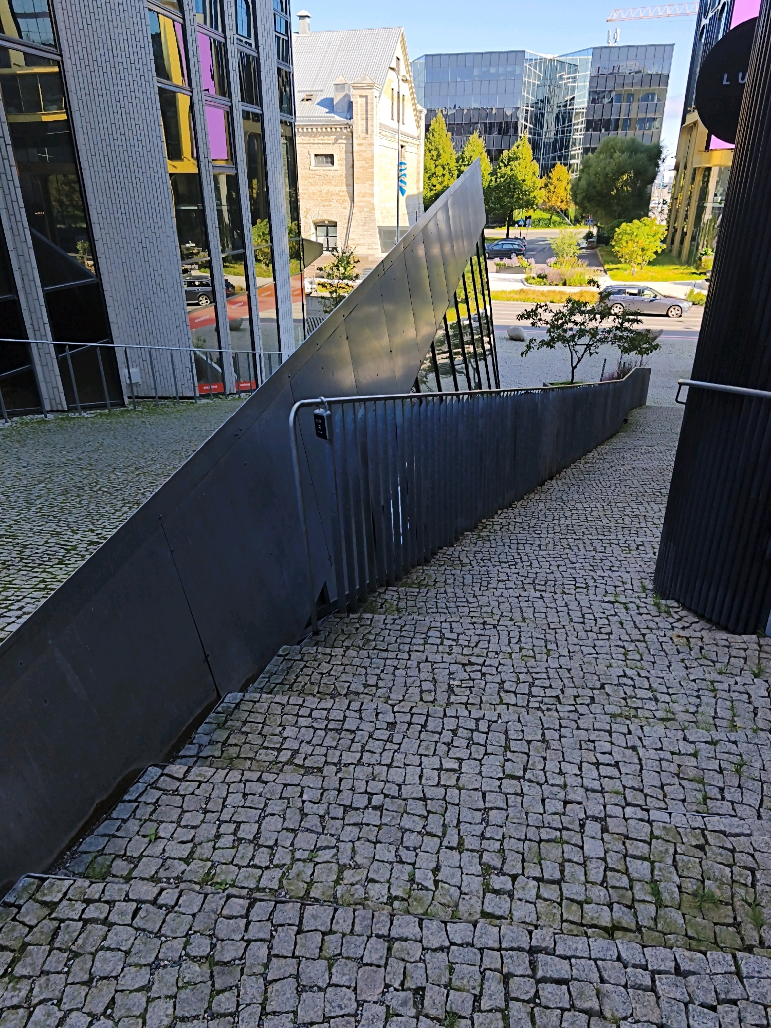 The image shows a modern architectural scene in Tallinn, Estonia, likely in a business district. The foreground features a pathway paved with cobblestones, descending from right to left. Along the left side of the path is a steep, dark metallic ramp-like structure, possibly an art installation or an architectural feature. A metal railing runs along the edge of this structure. To the left of the ramp, a glass-fronted building with a brick-like facade is visible, reflecting the surroundings, including trees and other buildings.
In the midground, a street is visible with cars driving. Beyond the street, several modern glass-and-steel office buildings are prominent, with trees showcasing autumn colors. The background includes a light-colored traditional building with a grey roof, and in the far distance, a construction crane can be seen. The sky is clear and blue. The scene is devoid of people. The overall impression is one of contemporary urban development integrated with some older architectural elements.