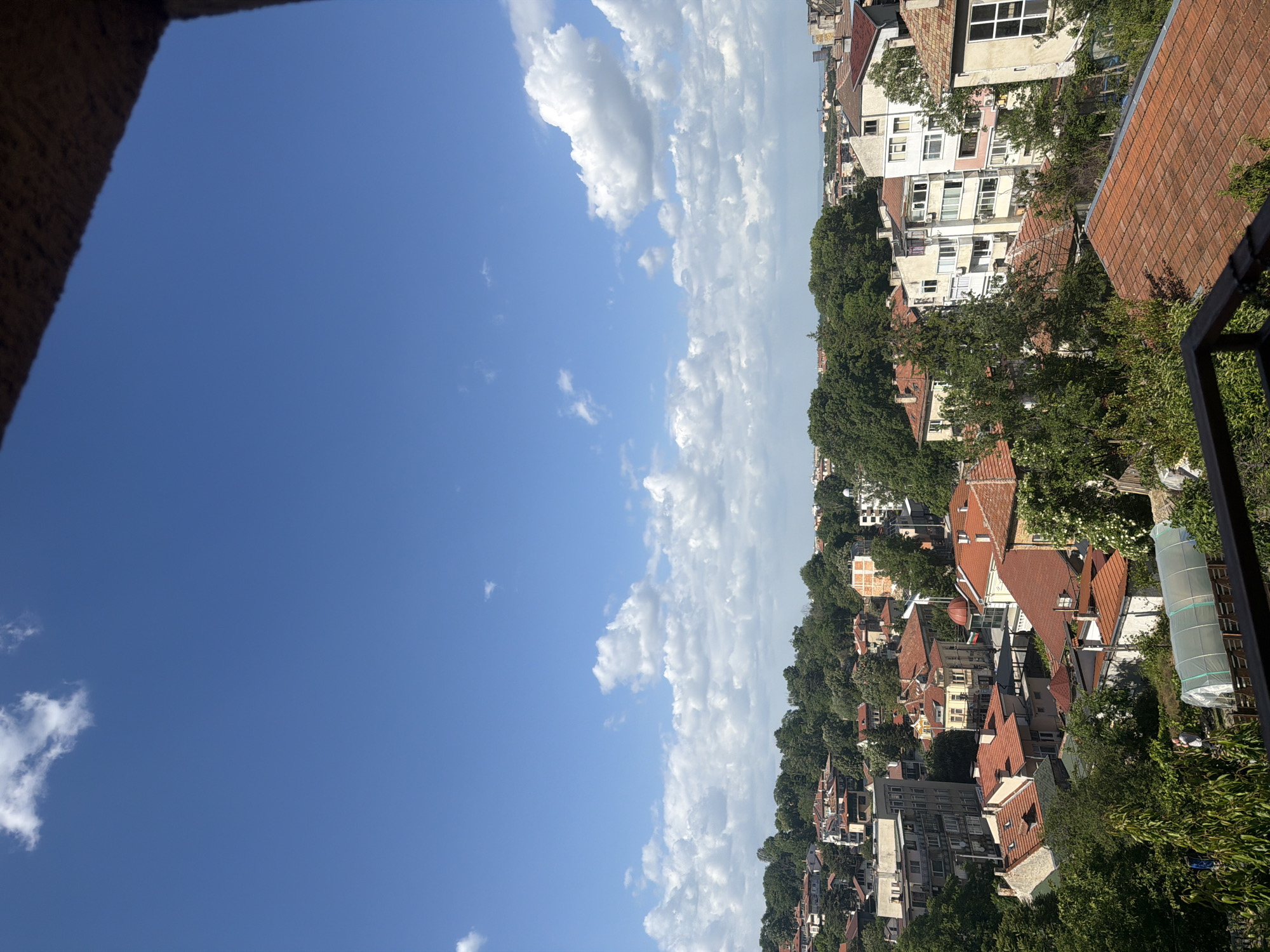 The image presents a high-angle view of Shumen, Bulgaria, under a blue sky with scattered clouds. The scene is divided; the left side shows the sky while the right displays a densely populated area with buildings, predominantly houses with red-tiled roofs interspersed with trees. Taller apartment buildings are also visible. Some foliage obscures parts of the view. The perspective is from an elevated position, possibly a balcony or higher floor of a building.