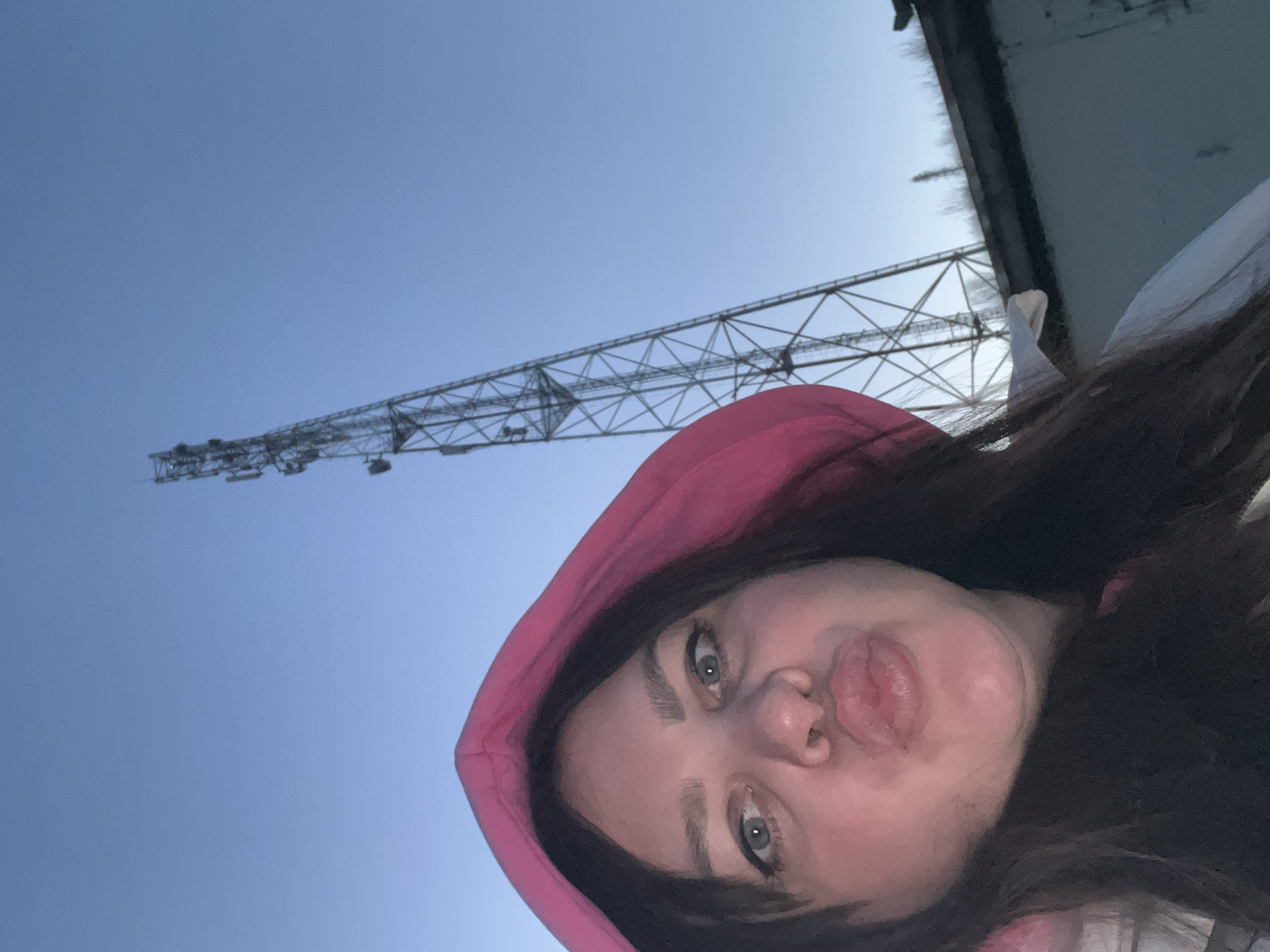 A selfie photograph shows a woman with brown hair wearing a pink hooded top against a blue sky. A tall metal structure, possibly a transmission tower, is visible in the background. The photo was taken at a low angle, showing the woman's face with her lips pursed. The location is Noviy, Russia.