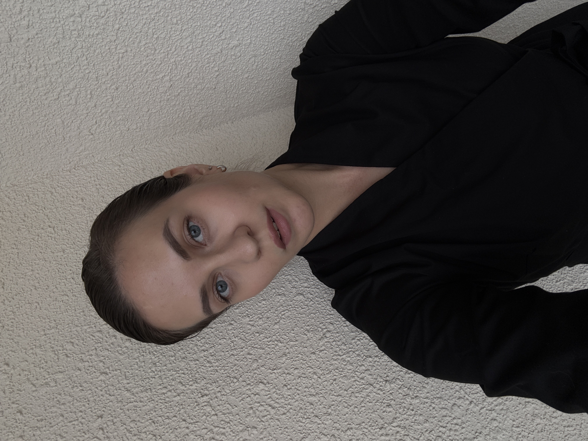 A person with light skin, light eyes, and dark hair slicked back is lying on a textured, light-colored surface. The person is wearing a black garment, possibly a shirt or jacket. The background appears to be a close-up view of a wall or similar surface. The location is not indicated in the image.
