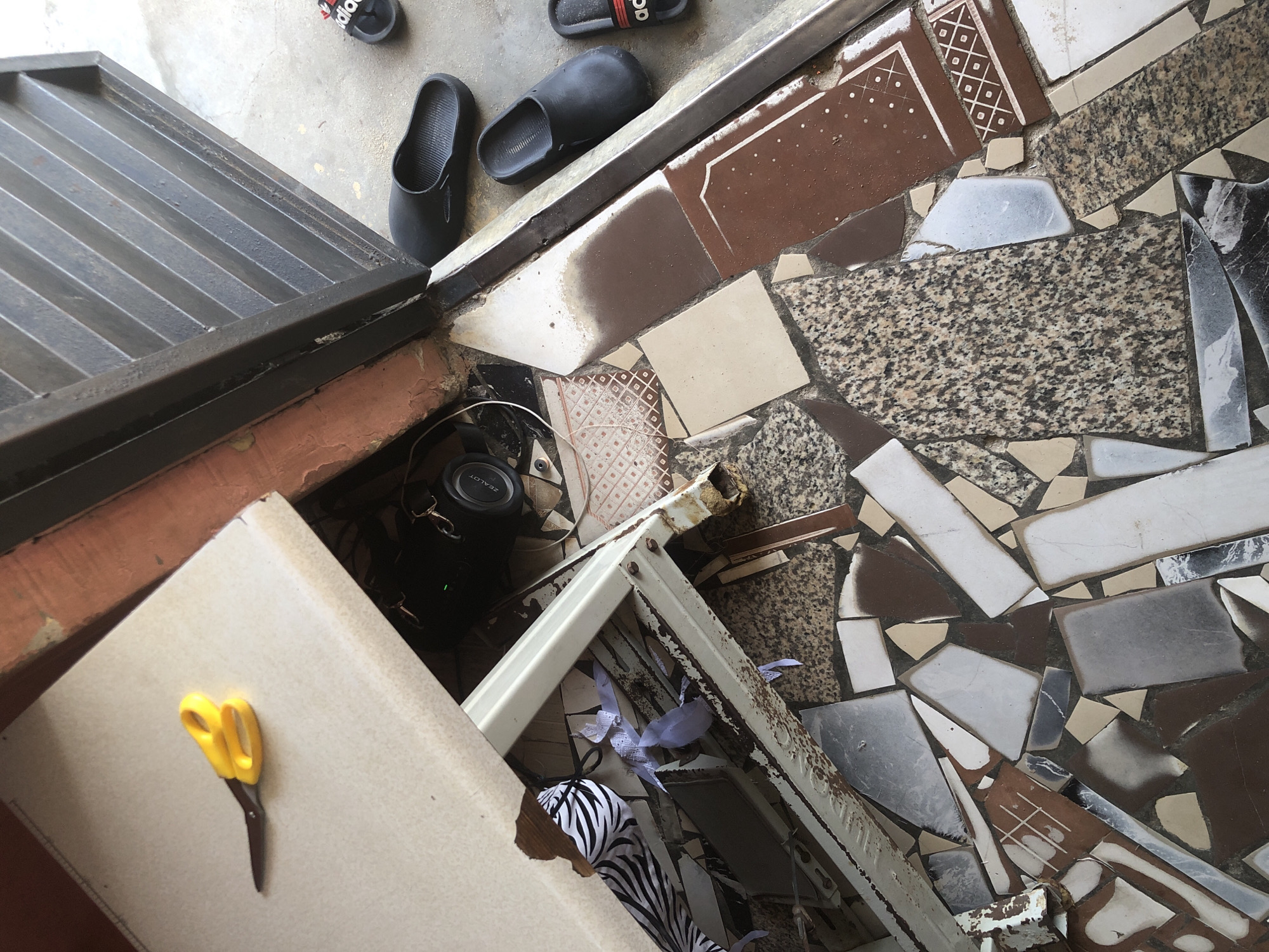 A pair of yellow-handled scissors lies on a light-colored, flat surface, possibly a table or shelf, in Ado, Nigeria. To the right of the scissors, a black portable speaker is visible. In the background, a black metal gate with horizontal slats is closed, and a concrete floor is visible beneath it. Several pairs of black slip-on sandals are placed on the concrete floor. The floor itself is covered with broken tiles of various colors and patterns, creating a mosaic effect. Some of the tiles are brown, white, gray, and speckled granite. Torn white paper pieces are scattered on and around a white metal shelf, which is rusty and appears damaged. A black and white striped fabric is also partially visible under the shelf. The scene appears to be outdoors, near an entrance or doorway.