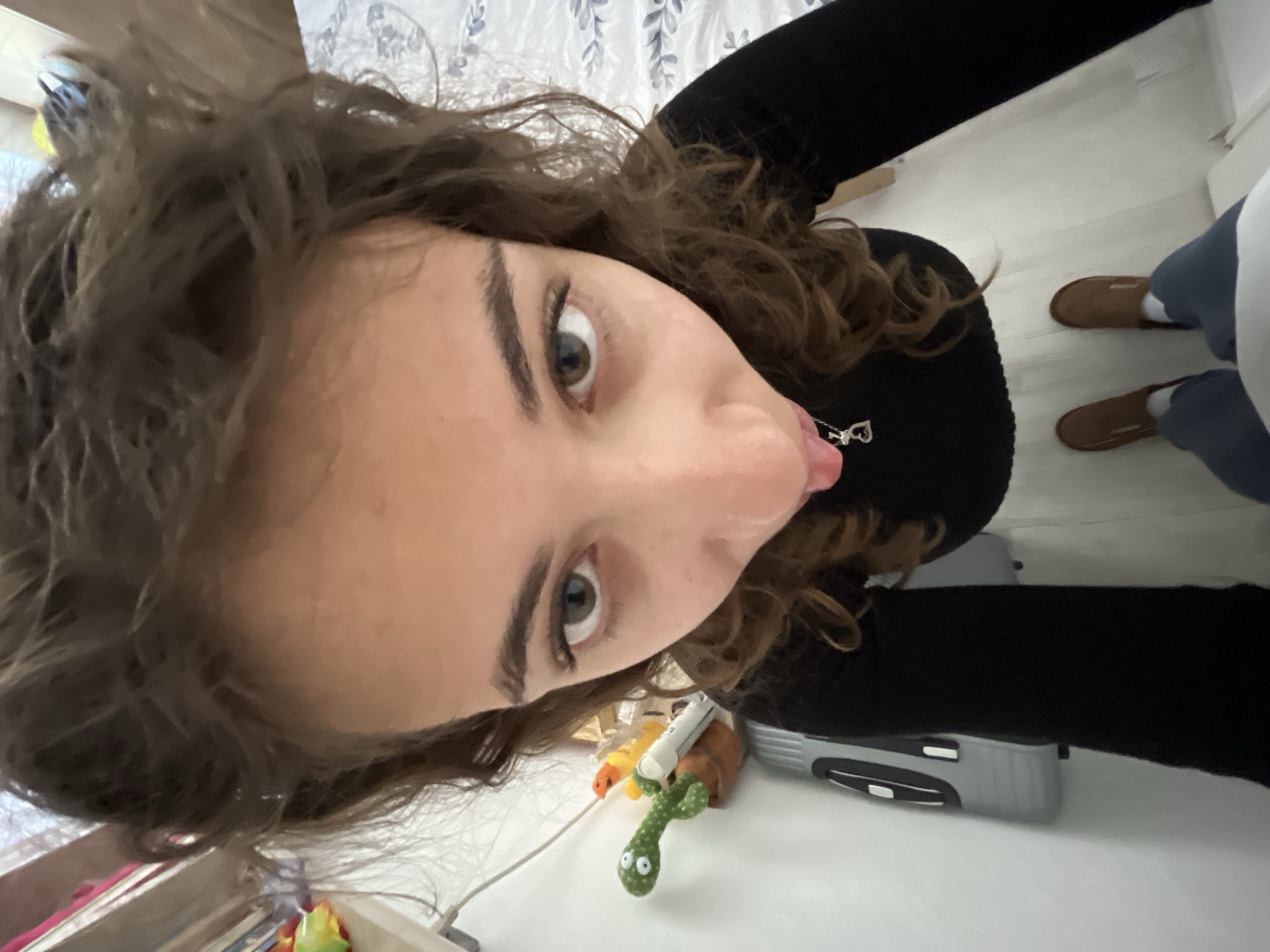 A close-up, distorted, high-angle selfie of a young woman with wavy brown hair, fair skin, and grey eyes, with her tongue slightly protruding. She is wearing a black long-sleeved top and a heart-shaped pendant. The background shows a cluttered white surface with various items, including a plush cactus toy, a gray-and-black object resembling a tool or device, and other unidentifiable objects. In the upper right, a portion of a person's legs, clad in blue pants and brown slippers with white socks, is visible. The location is difficult to ascertain from the image and cannot be confirmed as Karavelovo, Bulgaria.