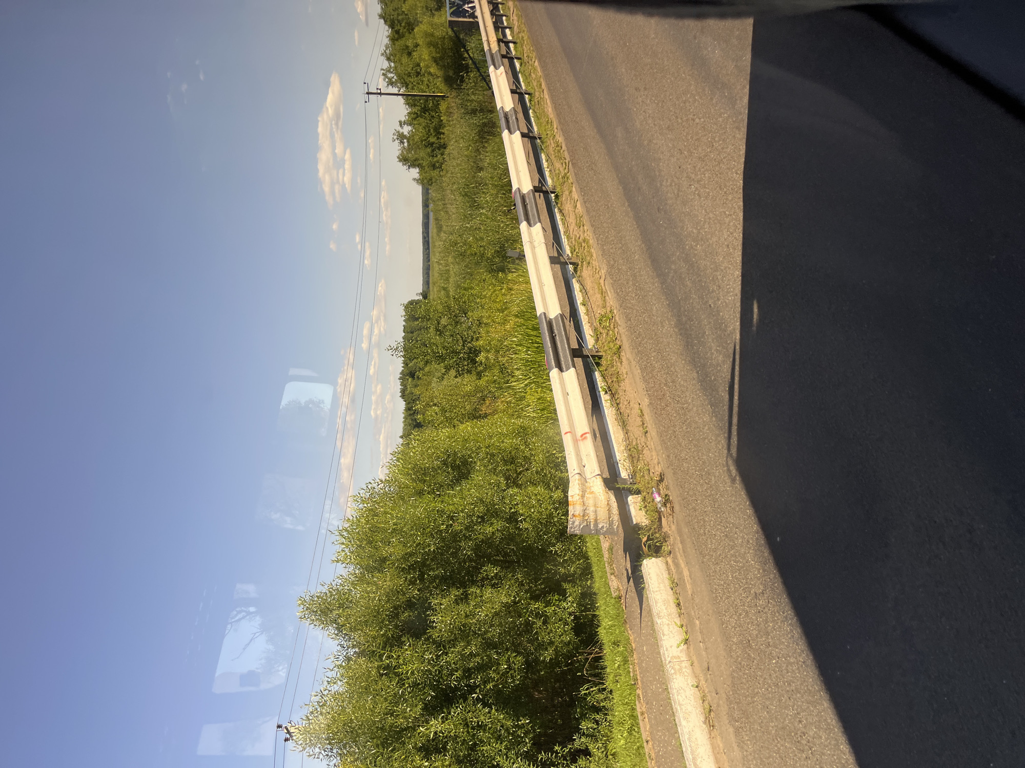 An asphalt road runs diagonally across the frame from the bottom left to the upper right. A guardrail with black and white stripes borders the right side of the road. Beyond the guardrail, there is a grassy area, followed by a dense stand of green trees and tall grass. The sky is a clear blue with a few scattered white clouds. In the distance, a body of water or a field can be seen. The scene appears to be a roadside view in a rural area, possibly near Myadzyel, Belarus, given the presence of deciduous trees and a landscape typical of Eastern Europe.