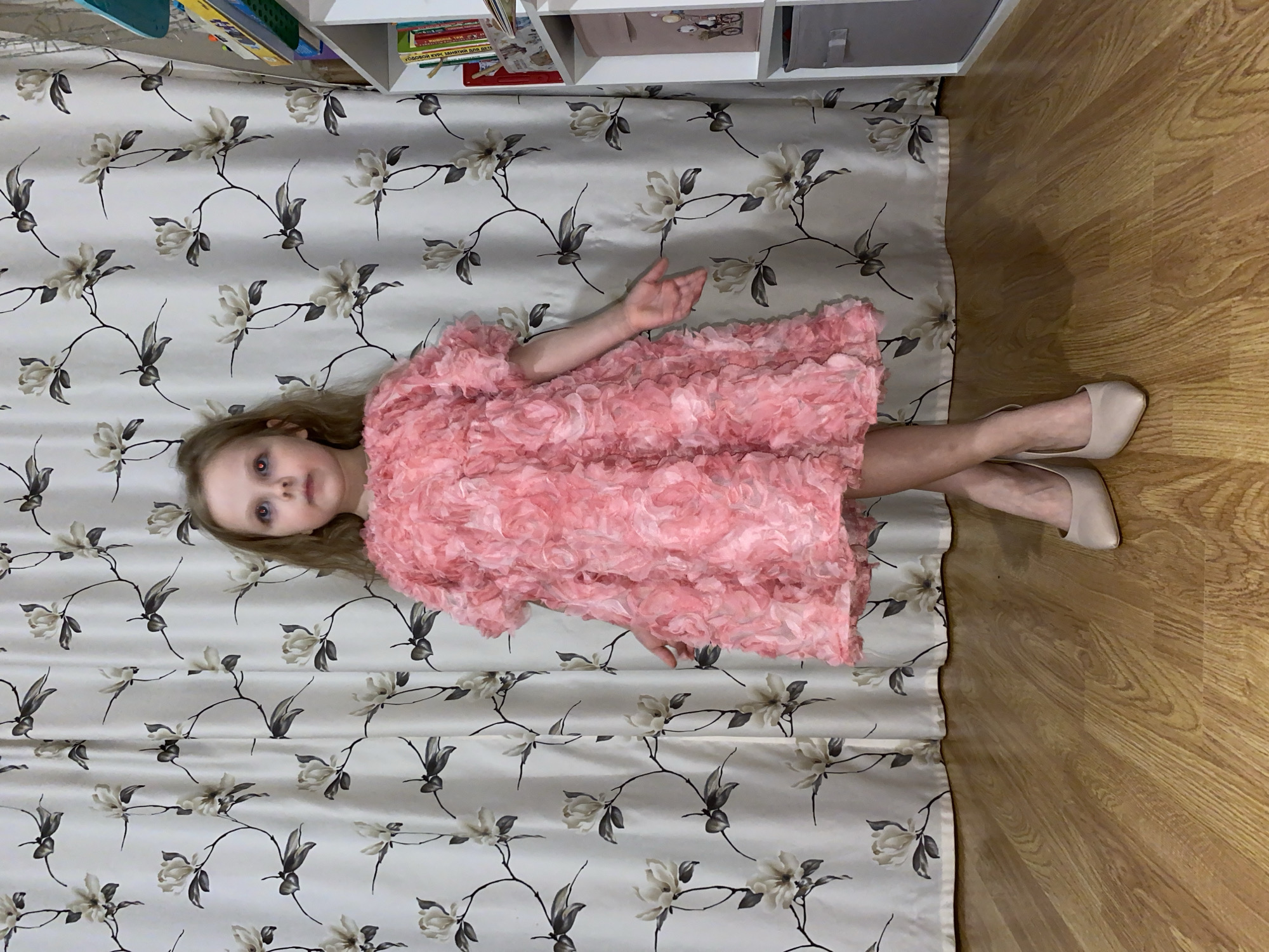 A young girl with long, light-colored hair is depicted lying on her back, looking upwards towards the camera, in what appears to be an indoor setting in Votkinsk, Russia. She is dressed in a vibrant, frilly pink dress with a textured, almost floral or petal-like surface. Her feet are adorned with light beige, pointed-toe flat shoes. The girl's eyes show a red-eye effect, suggesting the use of a camera flash.
She is resting on a patterned fabric, likely a curtain or a floor covering, which features an elegant design of light gray and white flowers on dark, winding branches against a cream background. To her right, a section of light-toned wooden floor is visible. In the background, white shelving units hold various books and objects, indicating a domestic and possibly child-oriented environment. One book spine clearly shows Cyrillic text, partially legible as "ГОВОРОЙ КУРС ЗАНЯТИЙ ДЛЯ ДЕТЕЙ" (Speaking Course of Activities for Children), providing a direct cultural and linguistic cue consistent with the location. The overall impression is a posed portrait of a child in a festive outfit.