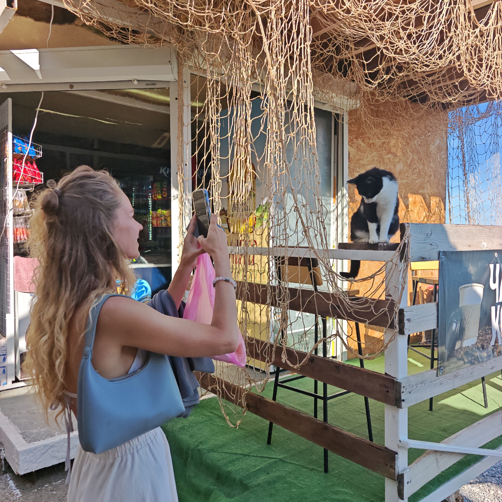 A young woman with blonde, curly hair is standing in front of a small shop or kiosk, taking a photo with her smartphone. She is wearing a light blue top and a white skirt, and she's holding a pink bag in her left hand. The shop has a rustic appearance with a wooden facade and fishing nets hanging as decoration. Behind her, a black and white cat is sitting on a wooden shelf, looking down.
The scene appears to be outdoors on a sunny day, suggested by the bright lighting and clear blue sky visible in the background. The woman's focus is on the cat, implying she is documenting her encounter with it. The setting looks like a coastal area or a tourist spot, possibly a cafe or souvenir shop, given the fishing net decor and the goods visible inside the shop. There's a sign visible on a fence behind the cat, but the text is not fully legible, though it appears to be in Cyrillic script, possibly containing the word "чай" (tea). The overall mood is casual and leisurely, capturing a moment of everyday interaction with an animal in a holiday-like setting.