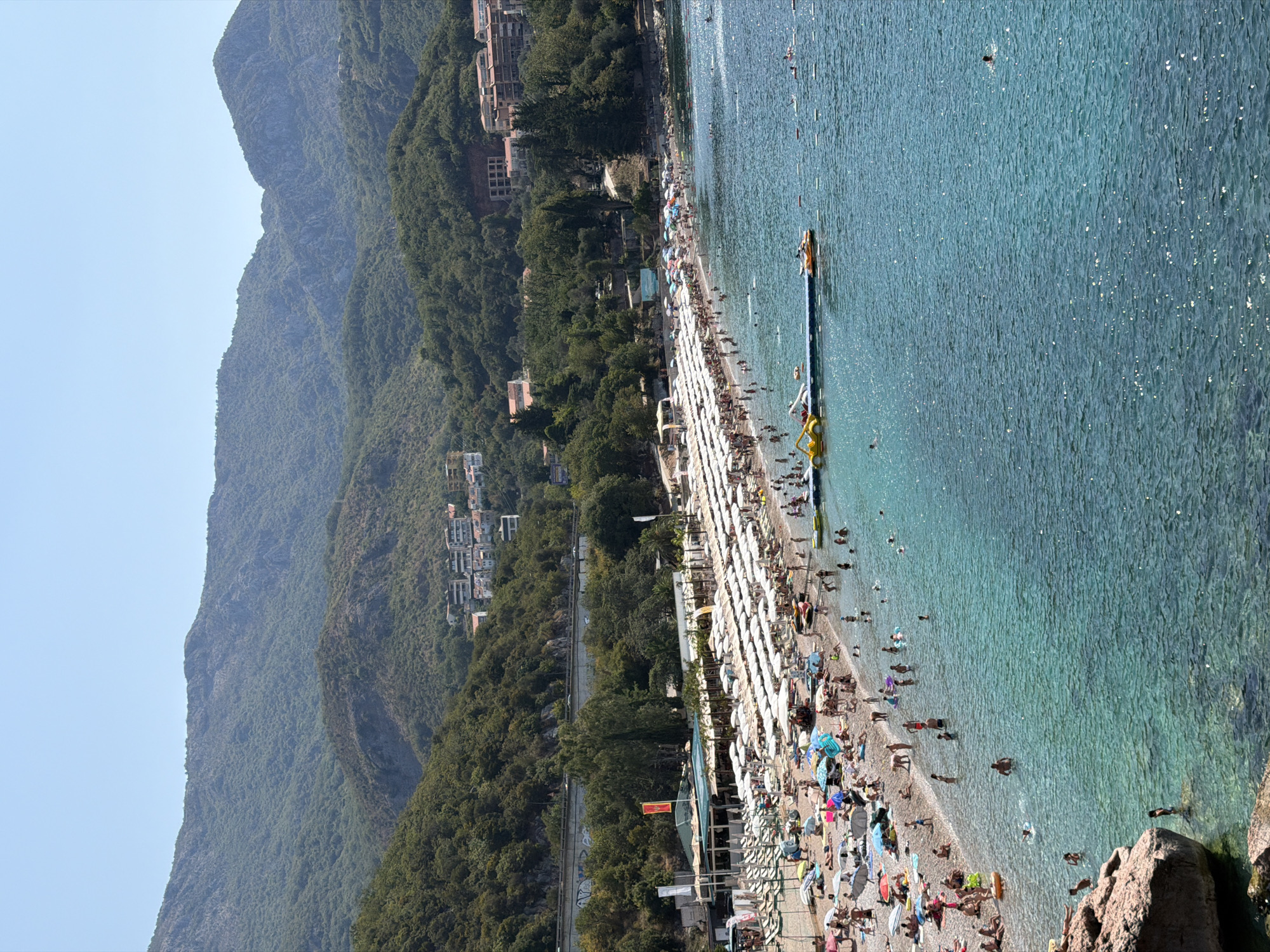 This image captures a vibrant beach scene in Bečići, Montenegro, during a sunny day. The foreground features a rocky outcrop jutting into the clear, turquoise Adriatic Sea. The main focus of the image is the expansive pebble beach, densely populated with sunbathers and lined with rows of white umbrellas and lounge chairs. Numerous people are visible on the beach, some relaxing under umbrellas, others walking along the shore, and children playing.
In the water, several individuals are swimming and wading, enjoying the refreshing sea. A floating water park, consisting of inflatable yellow structures, is anchored a short distance from the shore, with a jet ski nearby. A dark-colored pier extends into the water, equipped with seating and what appear to be more watercraft.
Behind the beach, a strip of greenery separates it from a road where cars are visible. Beyond the road, lush, green hills rise steeply, covered in dense vegetation. Buildings, including apartment complexes and hotels, are scattered across the lower slopes of the hills, indicating the presence of a developed tourist area. The sky above is clear and blue, suggesting fair weather. The overall atmosphere is one of a popular summer destination, with people actively engaging in beach activities and enjoying the coastal environment.