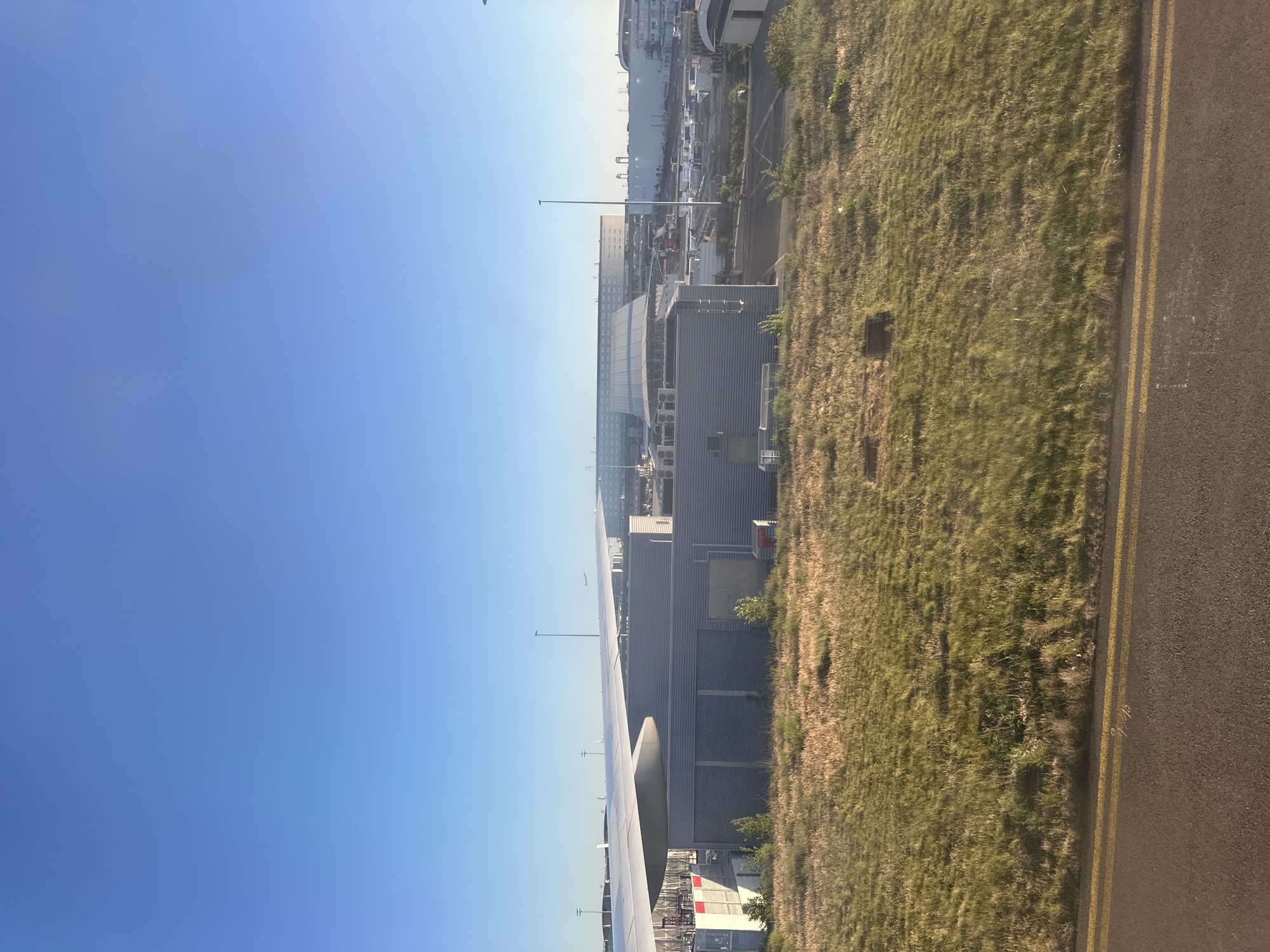 The image, taken from an airplane window, depicts an airport scene under a clear, bright blue sky. The perspective suggests the aircraft is on the ground, either taxiing or parked.
In the foreground, the robust wing of an airplane occupies the lower left portion of the frame, indicating the vantage point is from inside the cabin. The sky, a vibrant gradient of blue, fills the upper half, suggesting a sunny day with excellent visibility, likely morning or late afternoon given the quality of light.
To the right, a wide strip of grassy terrain, characteristic of airport peripheral areas, runs diagonally through the middle of the image. This natural barrier separates the paved surfaces from the main airport structures. Adjacent to the grass, on the far right, is a section of dark asphalt runway or tarmac, distinctly marked by two parallel yellow lines. Faint white numerical or textual markings, possibly "16" or "1/16", are discernible on the tarmac near these lines.
The middle ground and background reveal a bustling airport infrastructure. Numerous large buildings, likely terminals or hangars, are visible, featuring modern architecture with grey and reflective facades. A tall, slender pole or mast stands out against the horizon. Closer to the grassy area, smaller industrial-style buildings are also present. On one of these buildings, a red sign is visible with an illegible text, and another red sign displays a symbol resembling a fire extinguisher.
The setting appears to be a large, functional airport, consistent with the provided location context of Le Mesnil-Amelot, France, which is near Paris-Charles de Gaulle Airport (CDG). The scene is devoid of people, conveying a quiet moment at a busy transportation hub.