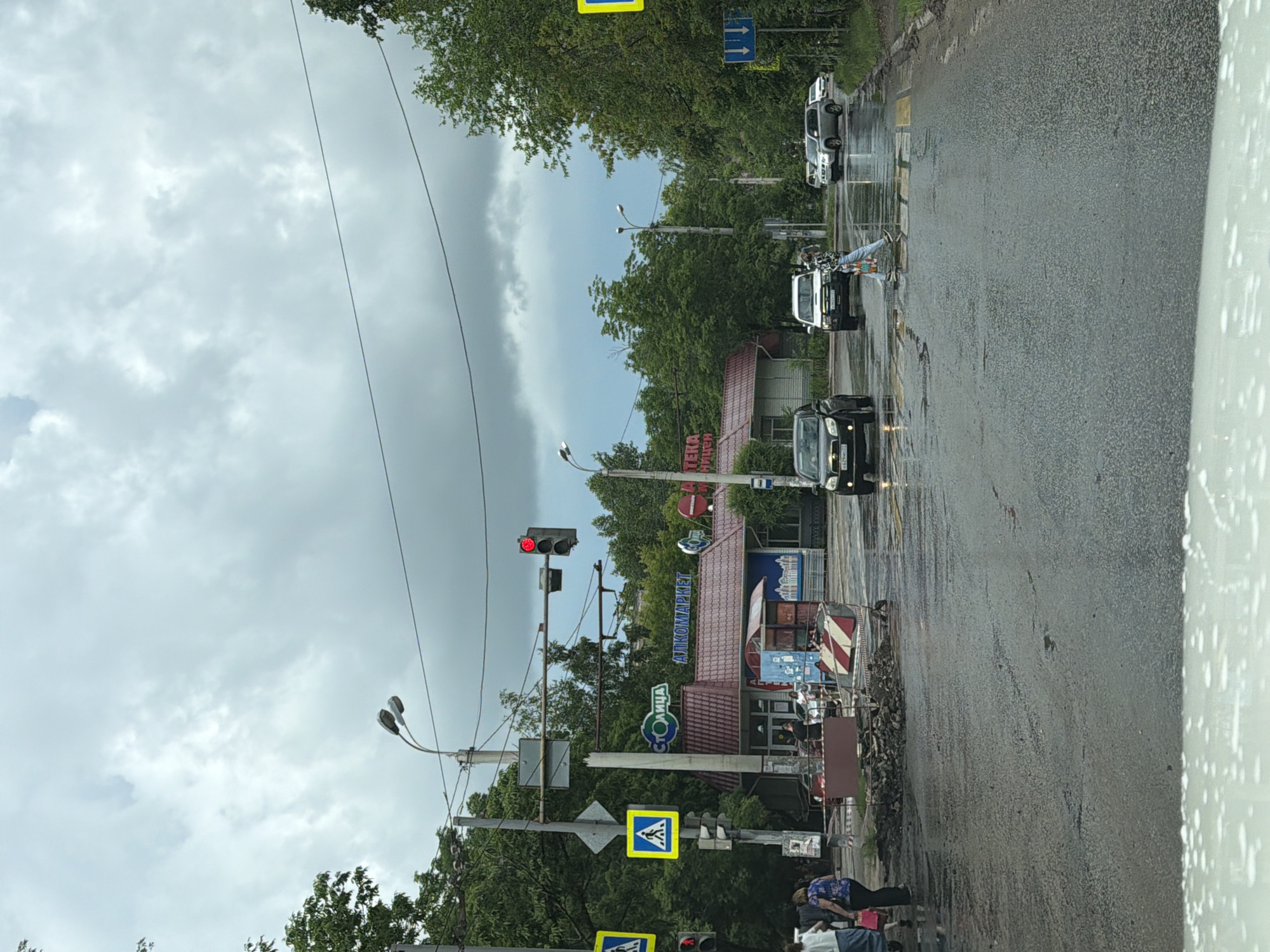 The image depicts a street scene in Khabarovsk, Russia, likely on a cloudy or rainy day, given the wet asphalt and overcast sky. In the foreground, the wet road surface is visible, with some scattered debris and construction barriers indicating ongoing work.
A crosswalk is present, with at least one person crossing. Several vehicles are on the road, including a black sedan, a white SUV, and a silver SUV. Traffic lights are visible, with one showing a red signal.
On the left side of the street, there is a building with signage that appears to be for a shop or market, with "СТОЛИЦА" and "АПТЕКА" visible, which translates to "Capital" and "Pharmacy." Pedestrians are visible near this building.
Further down the street, trees line the sides, and there are utility poles and wires. The overall atmosphere suggests a typical urban street environment in Khabarovsk.