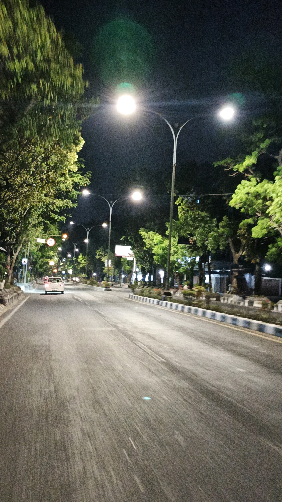 The image depicts a nighttime scene on a road in Surakarta, Indonesia, likely on Jalan Slamet Riyadi. The perspective is from the driver's seat of a vehicle, suggesting motion. Streetlights illuminate the asphalt road, which has visible lane markings. Trees with green foliage line both sides of the road, their leaves catching the artificial light. A white car is traveling away from the camera in the left lane. In the distance, more streetlights and trees are visible, along with some buildings and signs that are out of focus due to the motion blur. The overall atmosphere is one of a quiet, well-lit urban street at night.