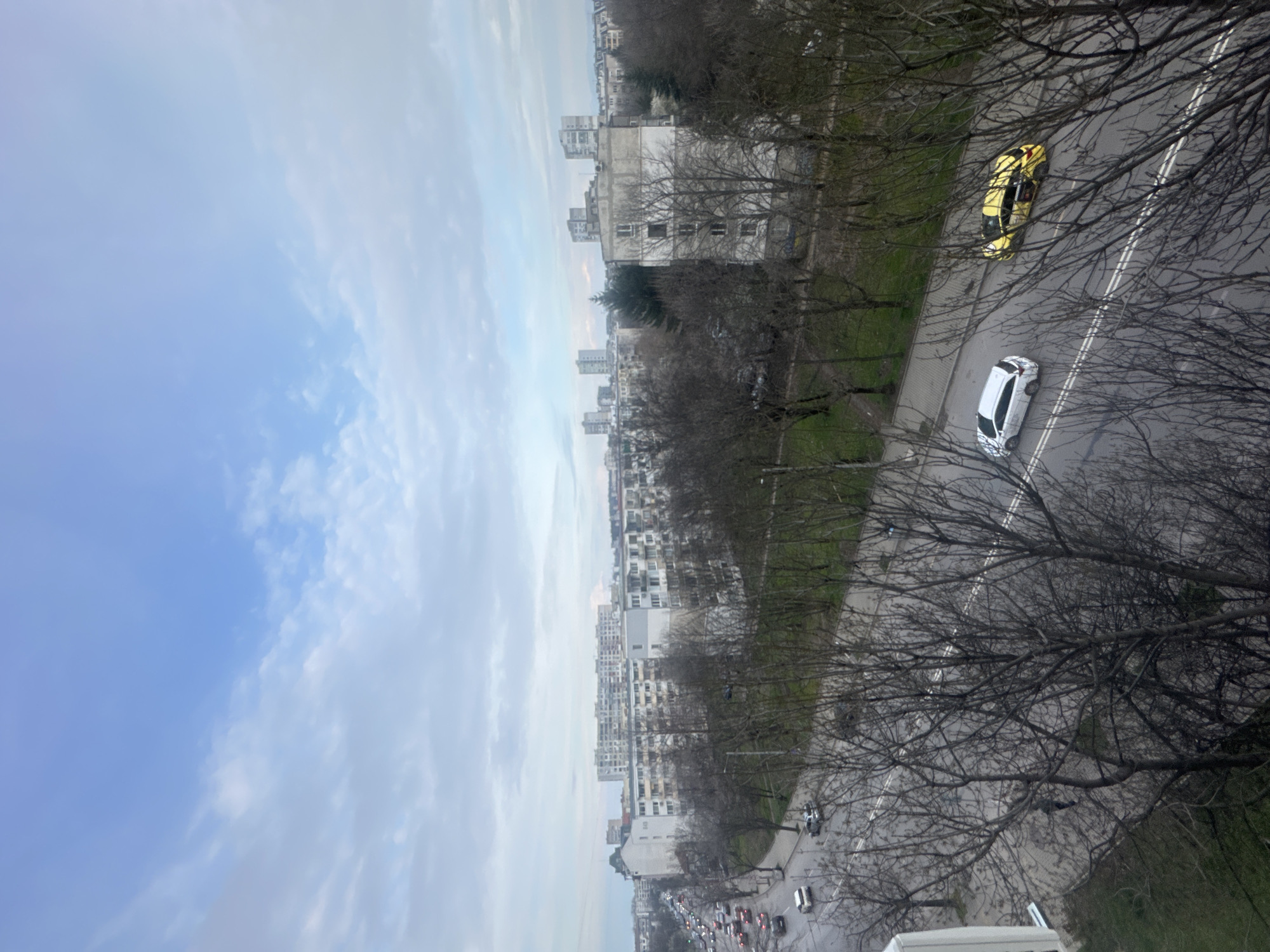 The image shows an aerial view of Sofia, Bulgaria. In the foreground, a road with sparse traffic is visible, with a yellow and a white car parked on the side. Bare trees line the road. Further in the background, dense apartment buildings stretch towards the horizon under a partly cloudy sky.