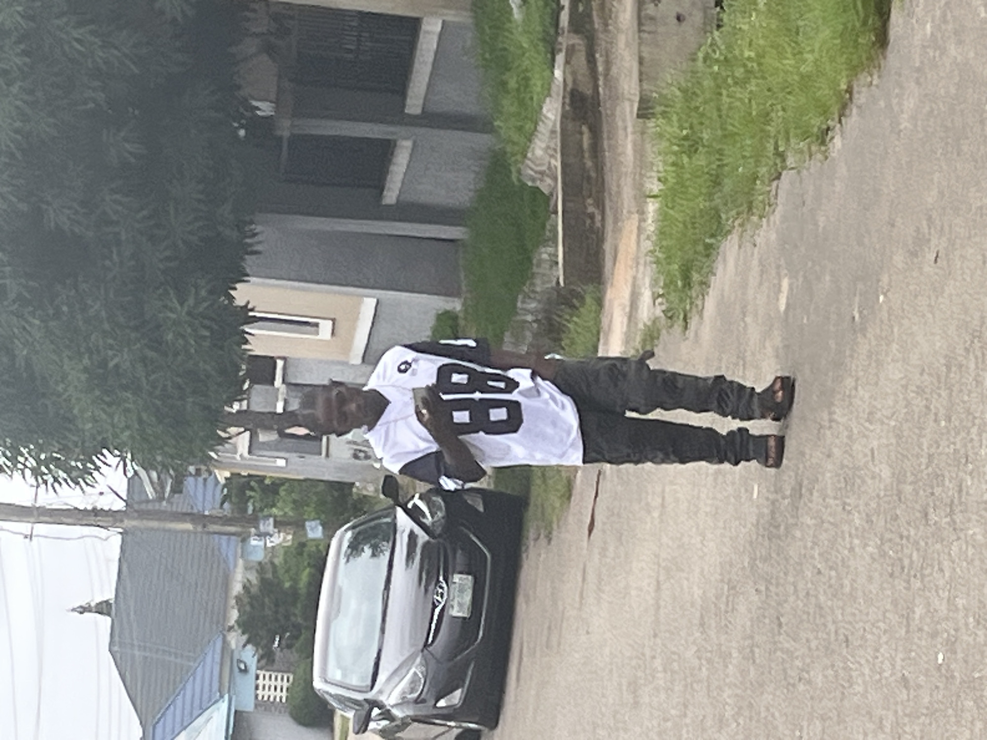 A young Black male wearing a white jersey with the number "88" on it and dark camouflage pants walks on a paved road in front of a dark sedan. He is also wearing sandals. The car is parked on the left side of the road. A two-story house with a gray facade and blue trim is visible behind the car. To the right of the house, there is a tall, leafy tree. The scene appears to be in a residential area of Abuja, Nigeria, indicated by the architecture and vegetation.