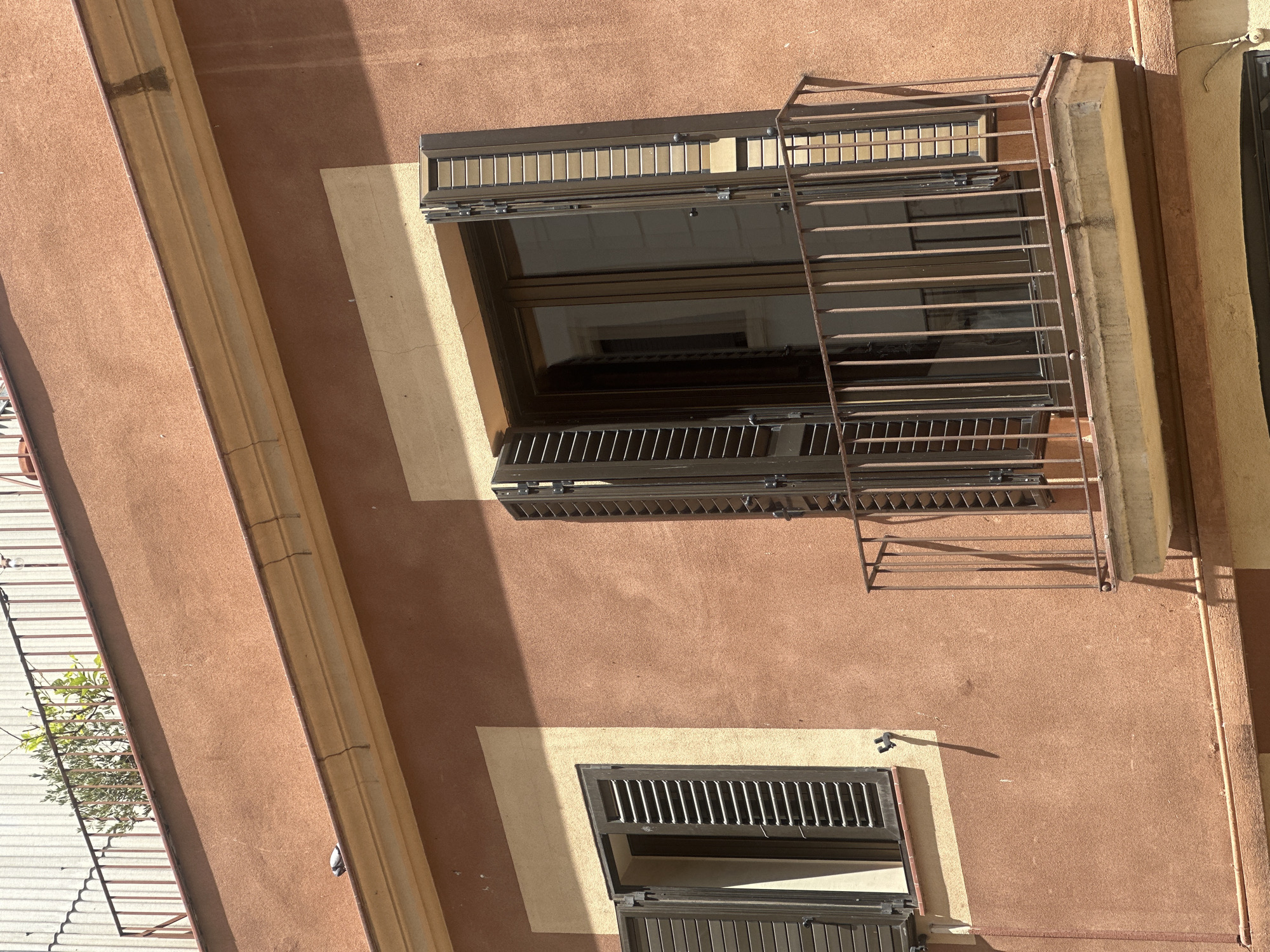 This is a photograph of a building's exterior, specifically focusing on two windows and a balcony. The building has a textured, orange-brown stucco facade. The upper part of the building features a light yellow trim and a darker orange stucco. A balcony with a rusted metal railing extends from the right side of the frame, in front of a set of double doors with dark brown shutters. The shutters on both windows are open and made of dark brown slats. The window on the left is smaller and has a similar dark brown shutter system. The building appears to be located in Barcelona, Spain, as suggested by the architectural style and the presence of a rooftop terrace with plants and a railing visible in the background, characteristic of the city's urban landscape. The lighting indicates a bright, sunny day.