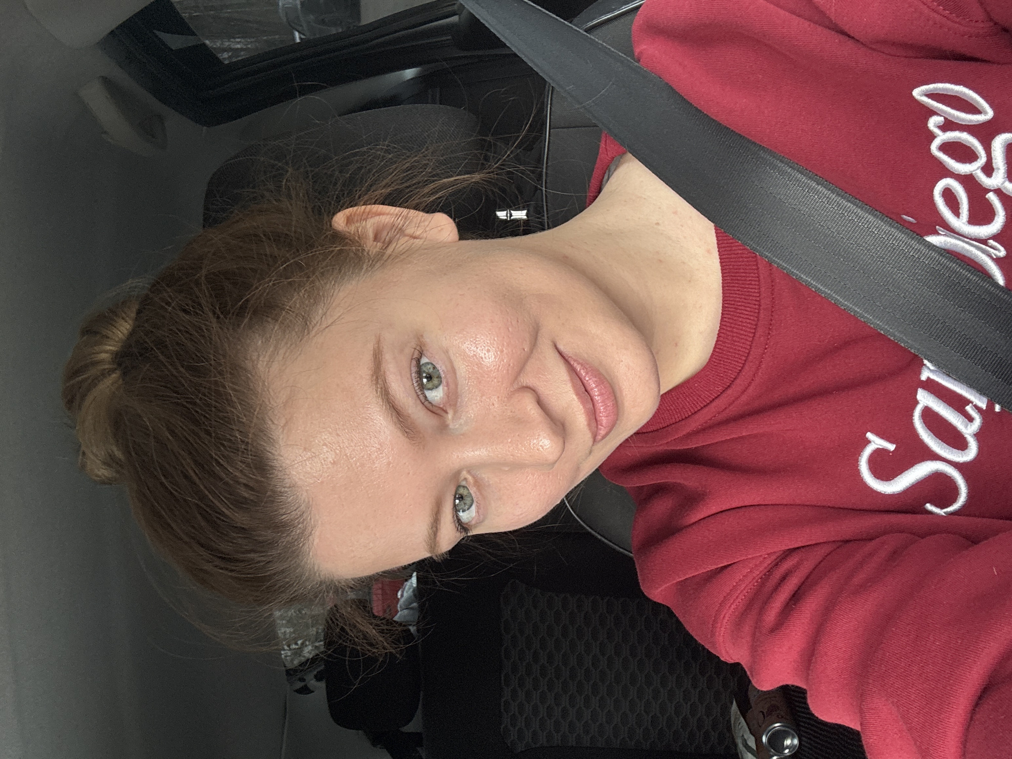 A woman is in a car, wearing a burgundy sweatshirt with white cursive writing "San Diego" on the chest. She is wearing a black seatbelt across her chest. Her hair is pulled back in a bun. She is looking at the camera. The interior of the car is visible, including the headrest, ceiling, and a glimpse of the car seat. The exact location within Chaikovsky, Russia, cannot be determined from the image.