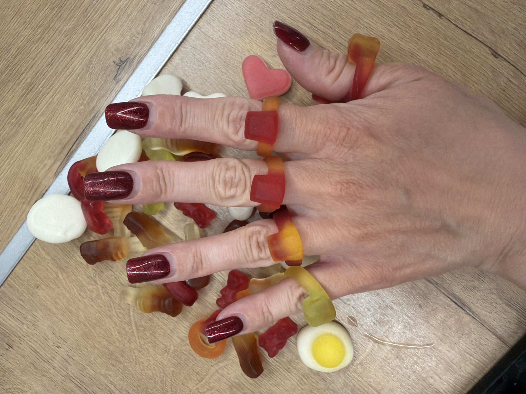 A human hand is positioned over a scattering of gummy candies on a wooden surface. The hand is adorned with five gummy rings, one on each finger. The fingernails are painted with a dark, sparkling red polish. The candies are of various shapes and colors, including hearts, eggs, and tubular forms. A silver strip borders the left edge of the wooden surface. The location and city of Votkinsk, Russia, are not evident in the image.