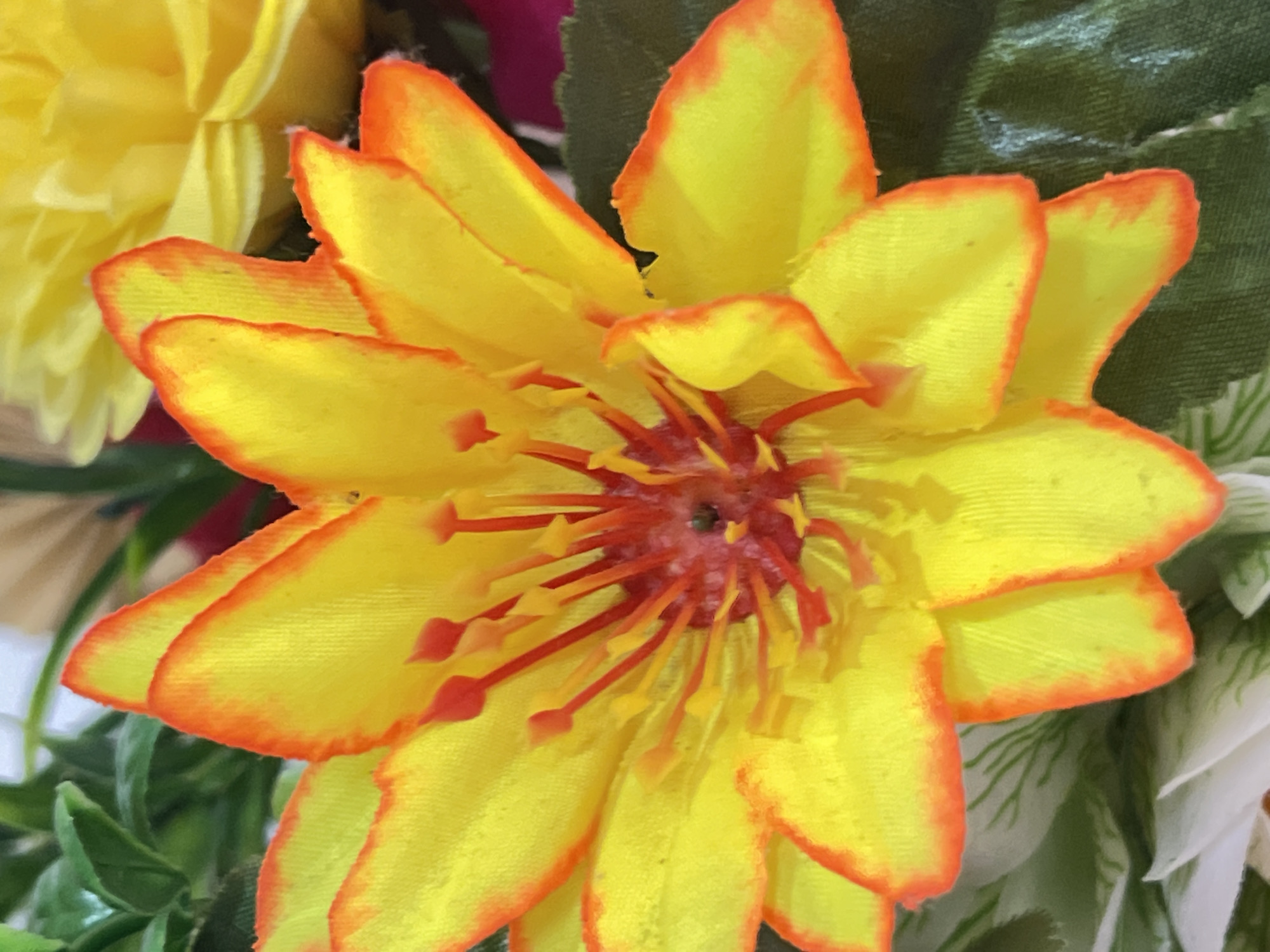 This is a close-up, eye-level, vertical, full-shot photograph of a vibrant yellow, artificial flower with bright orange edges. The flower has numerous petals radiating outwards from a central core. The center of the flower is a cluster of red stamens tipped with small orange anthers. The petals appear to be made of fabric and have a slightly textured surface. In the background, there are out-of-focus green leaves and a hint of another yellow flower. The lighting is bright, highlighting the colors and textures of the flower. There is no indication of the flower's location within the city of Denpasar, Indonesia, as the image focuses solely on the floral subject matter and provides no environmental context. There are no people or animals present in the image.