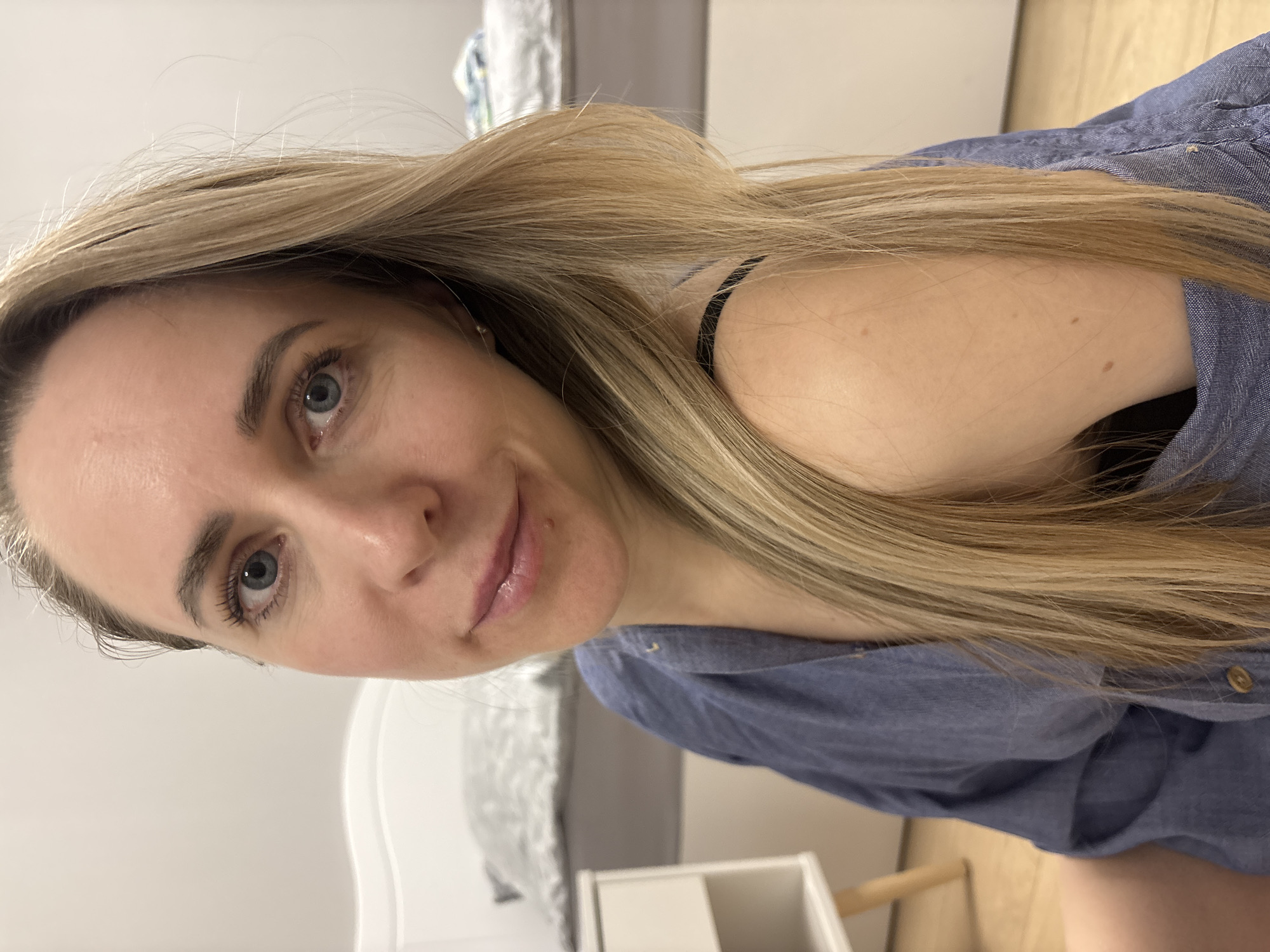 A woman with light blonde hair is indoors, wearing a blue collared shirt with a black strap visible under the shirt. Her face is in focus, showing light skin, blue eyes, and a slight smile. The background includes a white bed frame, white furniture, and light-colored wooden flooring. The location is not discernible from the image.
