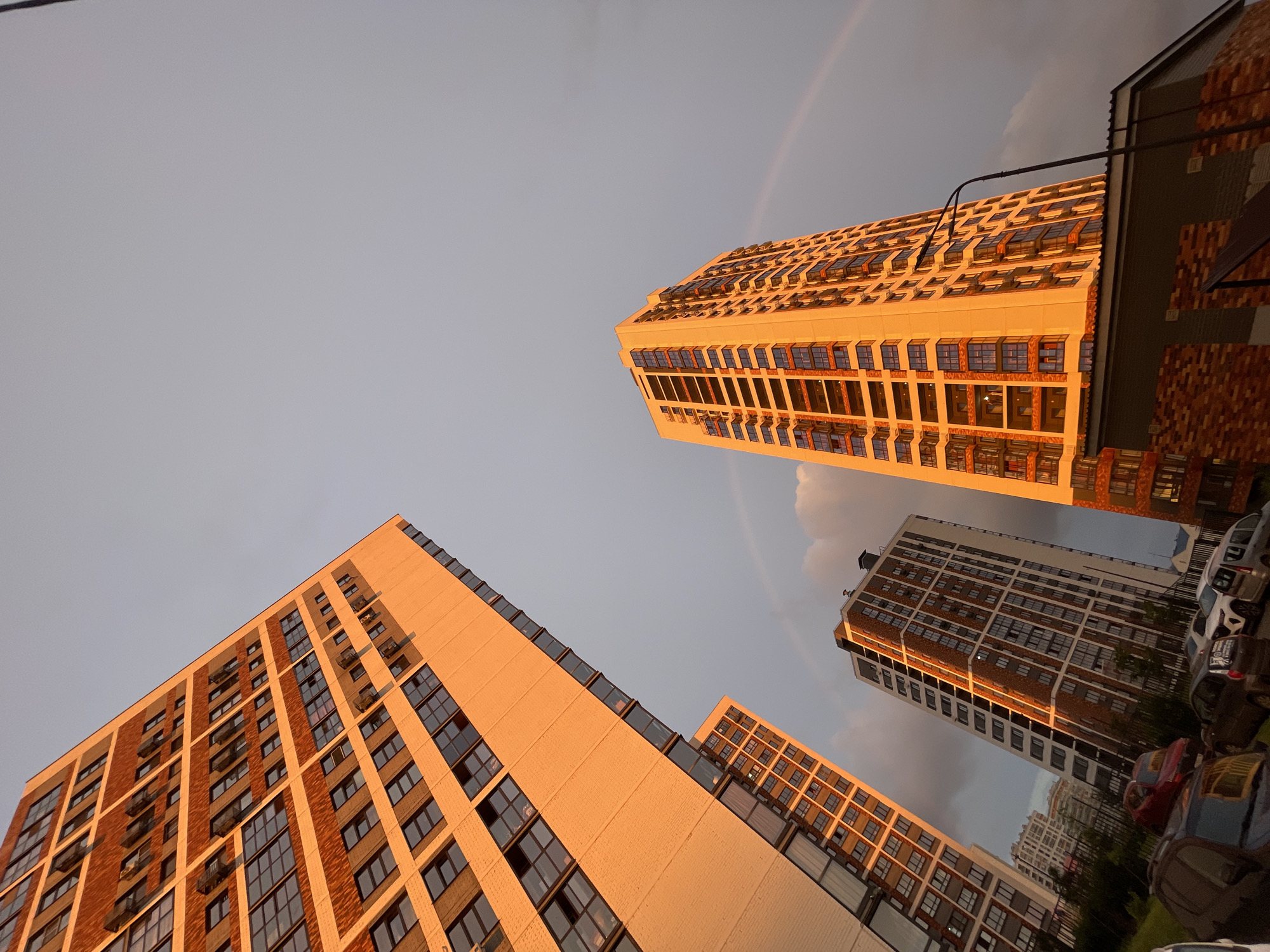 An upward-angled, full shot captures three modern, multistory apartment buildings illuminated by the warm, golden light of sunset. The buildings, characterized by their brick and glass facades, vary in height and architectural design, with balconies and numerous windows visible. A faint rainbow arcs across the sky in the background, with dark clouds juxtaposed against the bright sky. In the foreground, several cars are parked on a paved surface, and a streetlight stands tall to the right of the image. The overall scene suggests an urban residential area at dusk, with hints of recent weather, possibly a rainbow after a rain shower.