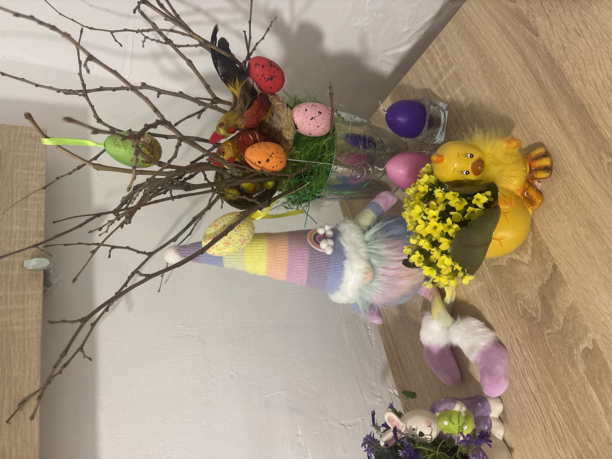 A still-life arrangement sits on a light wooden surface, likely a table, against a backdrop of white walls. The arrangement is Easter-themed.
The centerpiece is a branch arrangement with bare twigs. Colored Easter eggs are attached to the branches. There's also a faux bird attached to one branch. The twigs emerge from a clear glass container filled with green faux grass. Several colorful Easter eggs are within the grass.
Beside the glass container are two colorful Easter-themed candles. A plush Easter gnome with a pastel rainbow hat and white beard rests alongside the container. Adjacent to the gnome is a yellow Easter chick figurine holding a small potted plant with yellow flowers.
At the bottom of the frame is a decorative ceramic rabbit with painted eggs in a small garden of purple flowers.
Given the Easter theme, the location in Varna, Bulgaria is assumed to be a private residence and the arrangement is decorative.