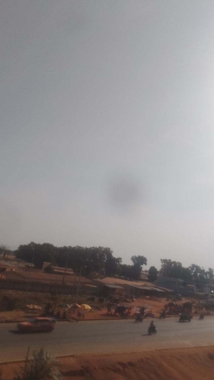 The image depicts an urban landscape in Korhogo, Côte d'Ivoire, under hazy atmospheric conditions. The upper two-thirds of the frame are dominated by a uniform, light grey, hazy sky, with a prominent dark, circular lens flare or smudge visible near the center.
The scene is viewed from a slightly elevated position. In the foreground, a reddish-brown unpaved embankment or dirt patch is visible, with some indistinct, blurred plant foliage at the very bottom.
A two-way asphalt road, appearing to have multiple lanes, occupies the lower third of the image, traversing horizontally. Traffic includes an orange-red car, likely a hatchback, blurred by motion as it travels left. Several motorcycles or mopeds are present; one is distinct in the right lane, moving right, while others are visible within the flow of traffic. A three-wheeled vehicle, possibly a cargo tricycle, is also on the far right, moving right.
Adjacent to the road, particularly on the right side, a bustling area suggests a market or roadside stalls. Multiple figures, indistinct due to distance and blur, are visible, along with structures partially covered by yellow and red tarpaulins or umbrellas.
Beyond the roadside activity, the terrain rises, characterized by reddish-brown earth and scattered low-rise buildings with dark roofs. Numerous trees with dark green foliage are distributed across the rising landscape, forming a denser line along the horizon. Several utility poles are visible vertically extending from the ground. The overall lighting is bright but diffuse, casting no distinct shadows, consistent with the hazy conditions.