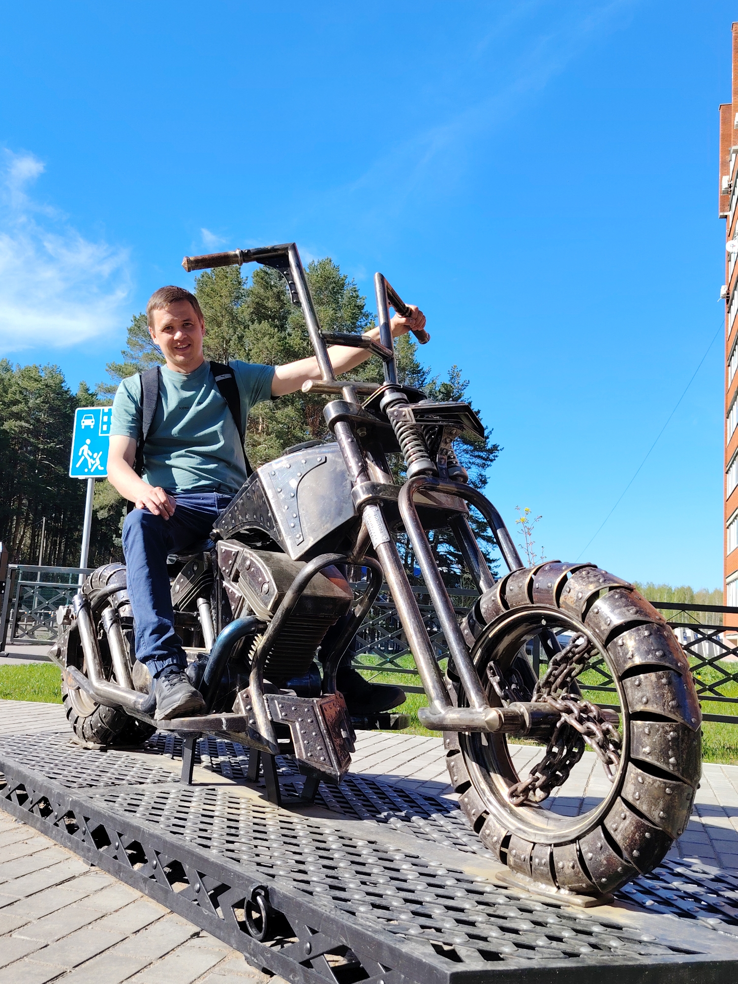 A full shot shows a man sitting on a metal sculpture of a motorcycle on an elevated platform in Chaikovsky, Russia.
The man, positioned on the motorcycle, looks directly at the camera with a slight smile. He is fair-skinned, with short brown hair, wearing a green t-shirt, dark blue jeans, and black sneakers. A black backpack rests on his shoulders. His left hand rests on his left thigh, and his right hand grips the motorcycle's right handlebar.
The motorcycle sculpture is constructed from various metal pieces, designed to resemble a chopper-style motorcycle. The wheels are made from a series of metal bands. A chain is present around the front wheel's axle. The bike is mounted on a black metal platform with a perforated surface.
Behind the motorcycle, there is a sign with a blue background, featuring a white symbol of a car and pedestrians. A fence runs along the background. Trees are visible behind the sign and fence. To the right, there is a building with a brick facade and white-framed windows.
The sky is bright blue with few clouds. The scene is well-lit, suggesting a sunny day. The location is Chaikovsky, Russia.