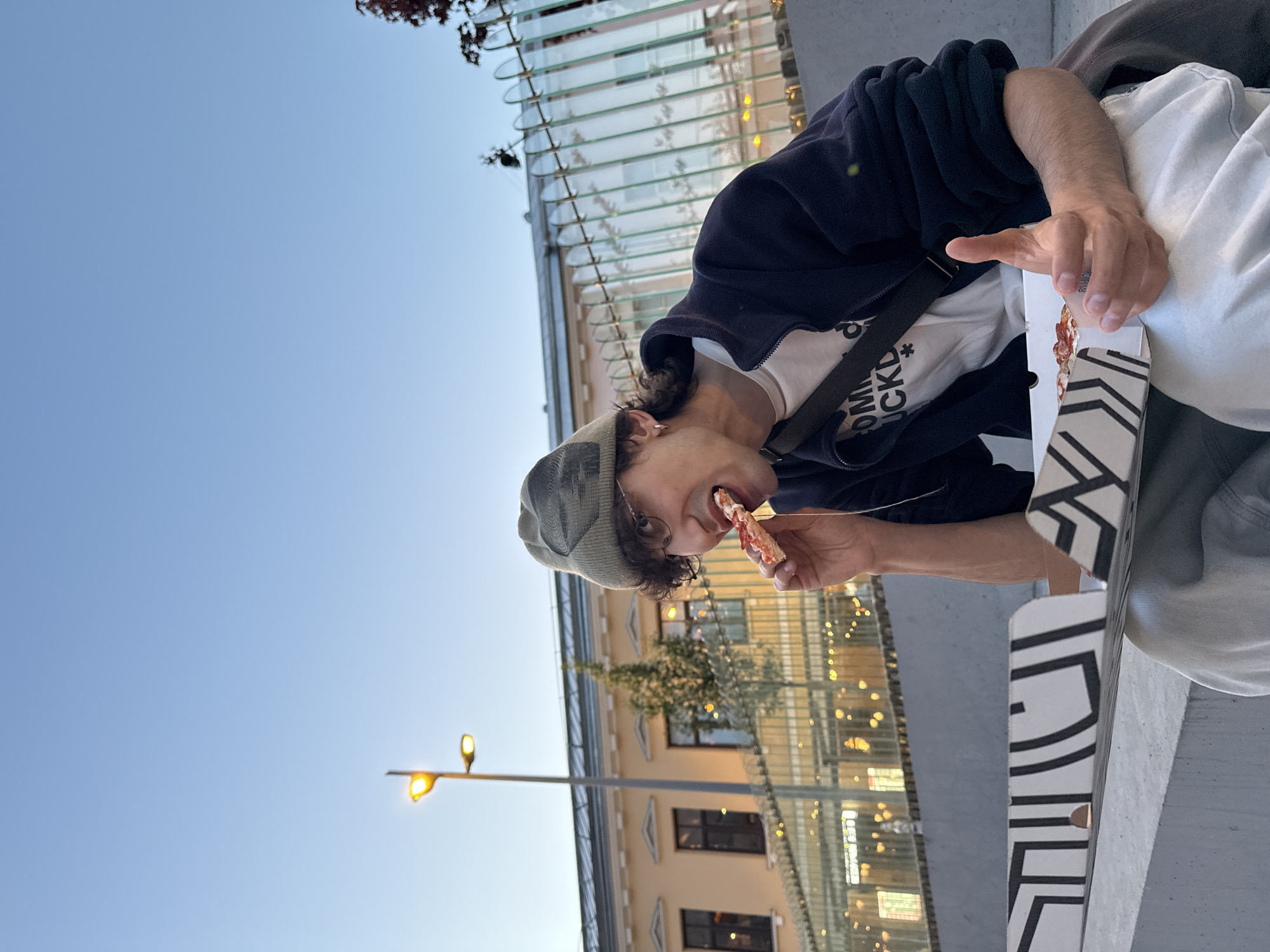 A person wearing a gray cap, a black jacket over a white T-shirt with printed text, and a black messenger bag strap across their chest is eating a slice of pizza. The pizza is from a white and black patterned pizza box. The person is holding the pizza slice with their right hand. In the background, there's a fence with a translucent roof, a building with lit windows, and a street lamp with two lights. The blue sky indicates it's likely daytime or dusk. The location appears to be in an urban environment, possibly Moscow, Russia, given the architectural style of the building and the street furniture.