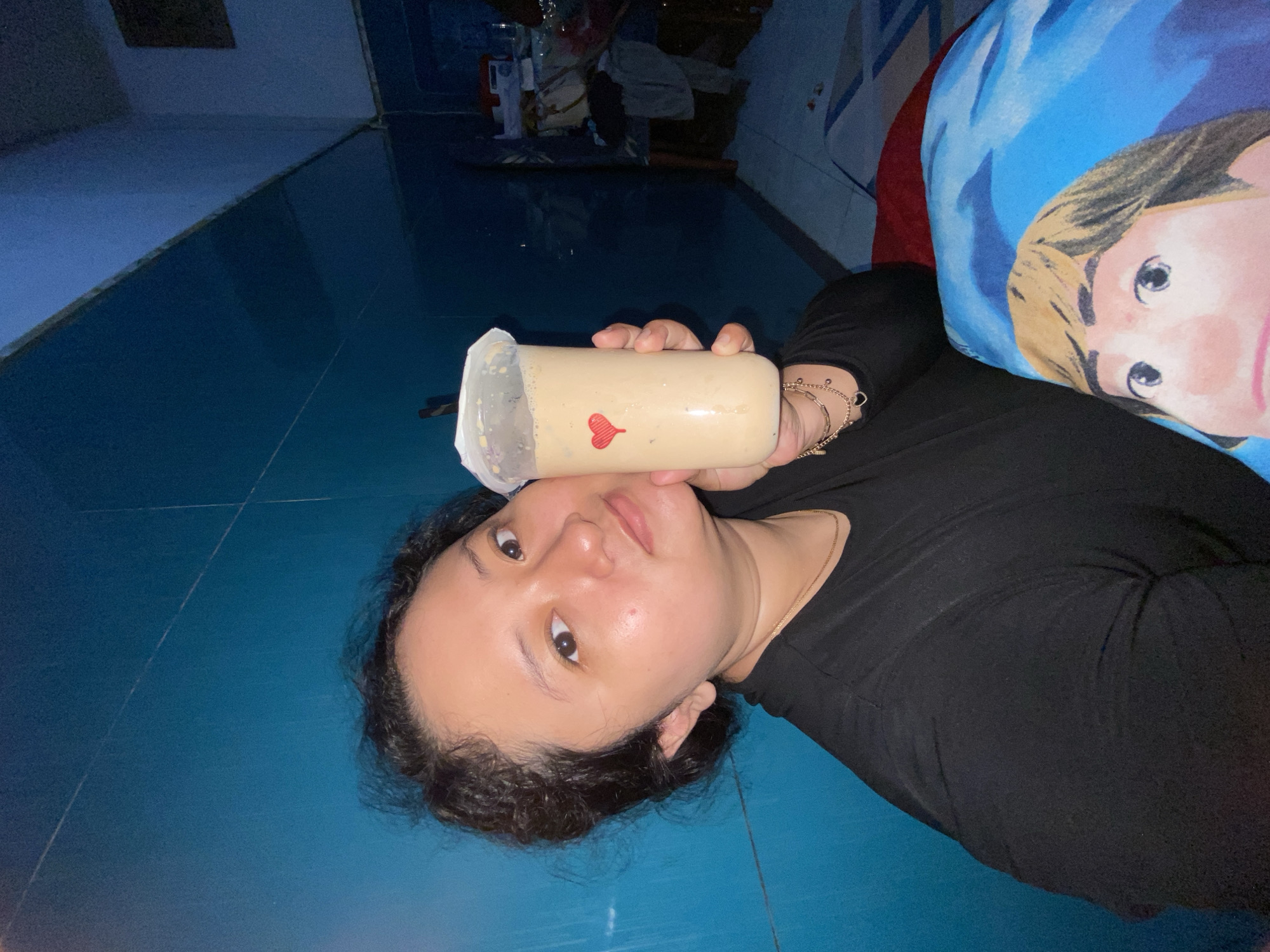 A woman is lying on her back on a blue-tiled floor in a dimly lit room. She is holding a plastic cup filled with a light brown liquid, adorned with a small red heart. The woman has dark curly hair, light skin, and is wearing a black top. Next to her is a blue pillow with a cartoon image on it. In the background, there is some clutter on a shelf or table. The image was taken in Gelam, Indonesia.