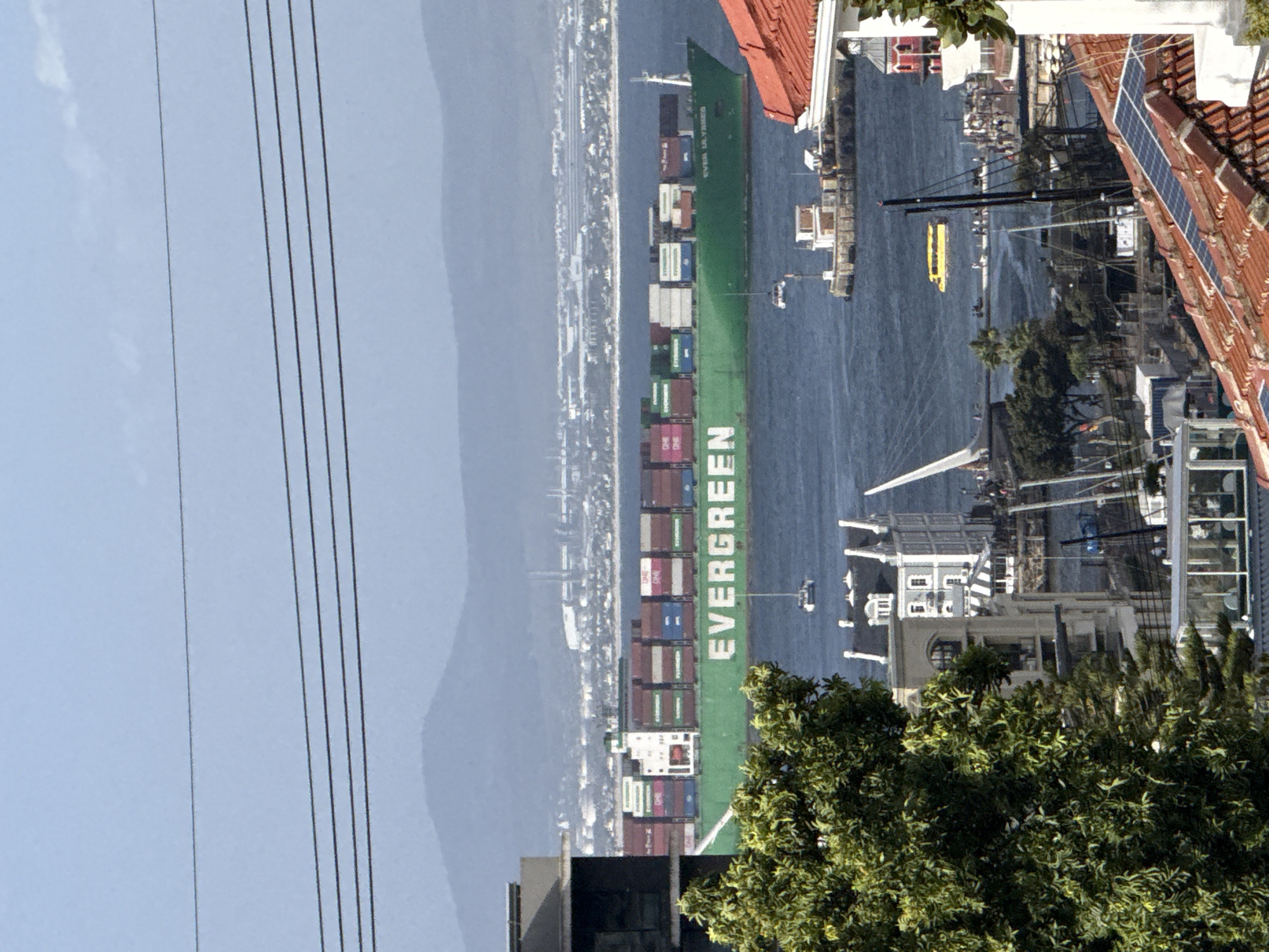 A container ship, labeled "EVERGREEN", is in the foreground of a harbor or bay area. The ship is stacked with shipping containers of various colors. On the right side of the frame, there are buildings with red tile roofs, and what appears to be solar panels on one roof. Trees are visible near the bottom of the frame, partially obscuring the view of the harbor and the waterfront. In the distance, across the water, is a city or town stretching along the shoreline. The day is clear with a blue sky, but there is some haze or fog over the mountains in the background. Power lines are present in the left side of the frame. There's a yellow boat also visible in the harbor. Buildings are also visible on the right side of the ship with some vegetation scattered around. The location appears to be in Cape Town, South Africa, based on the architecture, the geographical features, and the harbor setting.