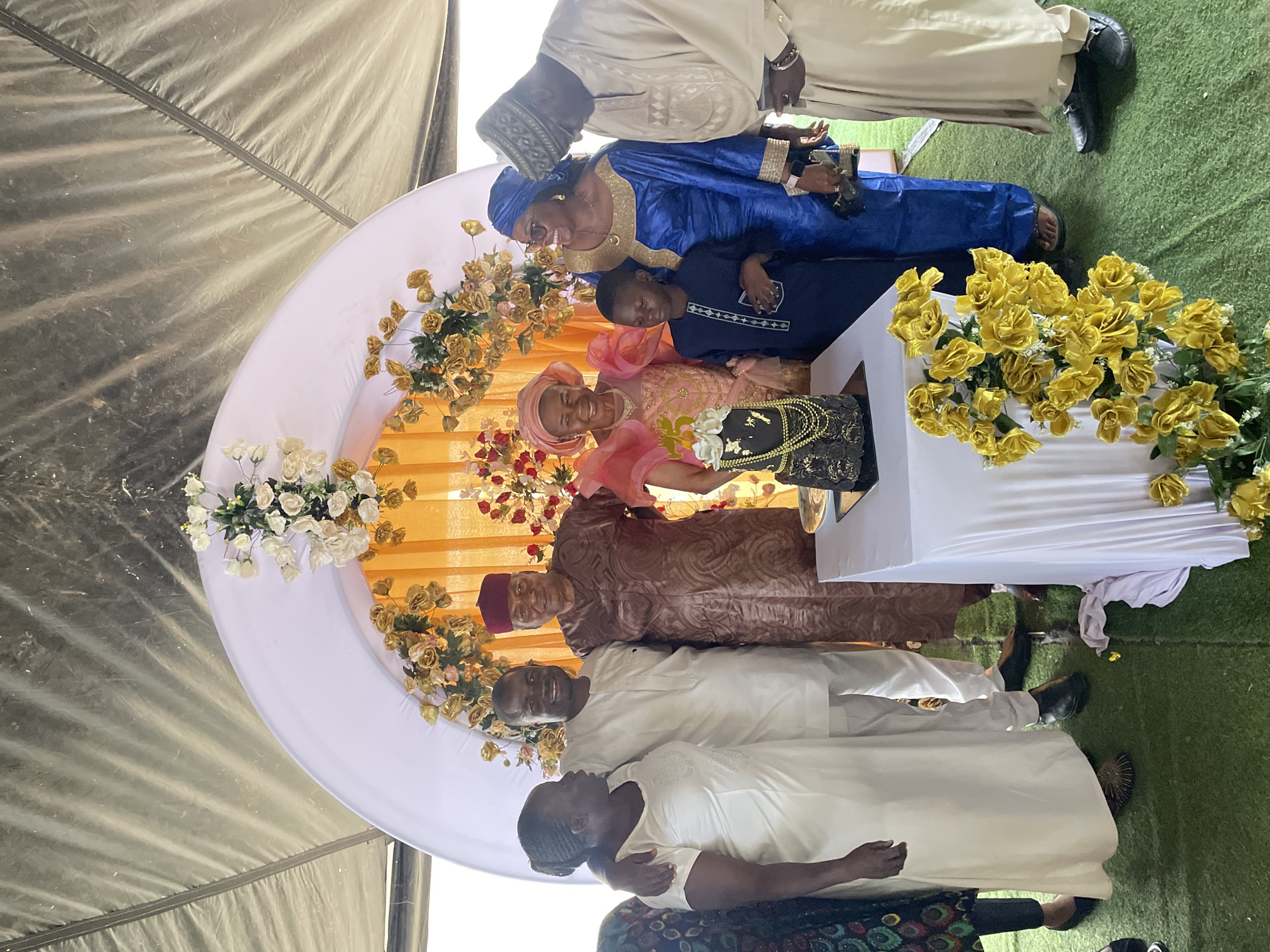 Here is a detailed description of the image:
**Scene:** A group photo is taken indoors, likely within a decorated tent or event space, against a backdrop adorned with white, yellow, and red flowers. The event seems celebratory.
**Location:** The scene is in Jos, Nigeria.
**People:**
* There are eight people in the photo. The people are dressed in ceremonial clothing.
* There is a woman in a pink headscarf and a dress holding flowers and smiling.
* There is a woman in blue clothing wearing a matching headscarf.
* There is a child wearing navy blue.
* There are a few men dressed in light clothing.
* The group is comprised of varying ages, from a child to older adults.
**Objects:**
* A decorative white semicircle serves as a backdrop.
* Flowers of various colors (white, yellow, red) are arranged around the backdrop.
* A table covered with a white cloth is present.
* A black purse is placed on top of the table.
* A piece of green material is on the ground.
**Actions and Interactions:**
* The group is posing for the photograph.
* Some individuals are smiling.
* The group appears to be in a celebratory gathering.
**Environment:**
* The event seems to be under a tent.
* The artificial grass on the ground.