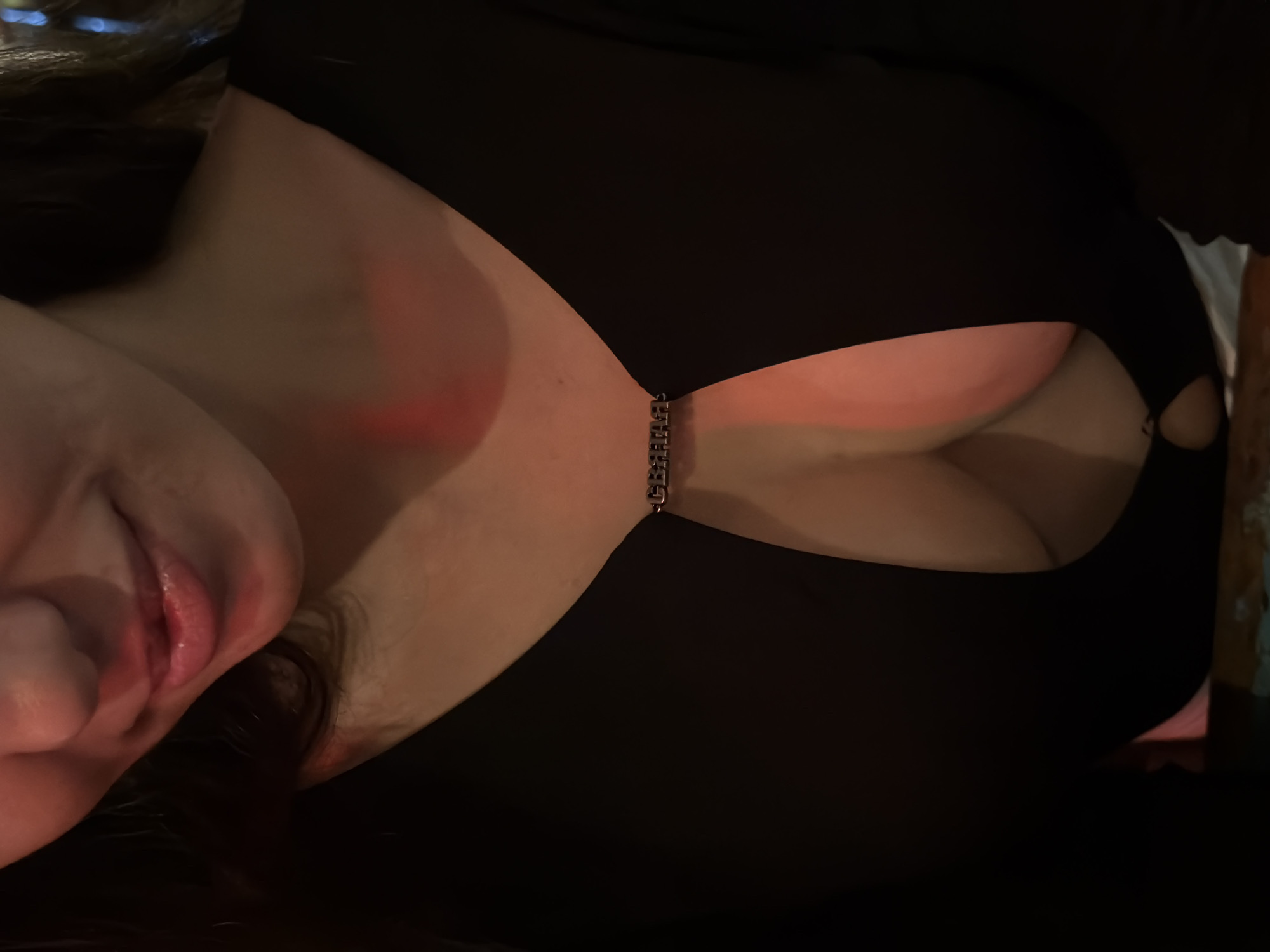 A rotated close-up captures a person, likely female, from the chest up, in what appears to be a dimly lit indoor setting in North Kuta, Indonesia. The individual's head is tilted, showcasing a partial view of her face, with prominent full lips exhibiting a slight smile. She wears a black garment, possibly a top or dress, featuring a deep V-neckline. This neckline is uniquely adorned and held together across the sternum by a short, metallic, silver-toned chain. Upon closer inspection, the text "VERSACE" is clearly visible on the links of this chain.
The skin on her neck and chest displays a noticeable reddish glow, indicative of ambient red or warm lighting in the environment. Dark hair is visible framing the side of her face. The black garment also includes a small, subtle circular cut-out design on the right side, near the armpit area. The background is largely dark and out of focus, with only vague blurs of light in the upper left corner and an indistinct object on the far right, further emphasizing the low-light conditions. The overall atmosphere suggests an intimate or relaxed evening or nighttime scene.