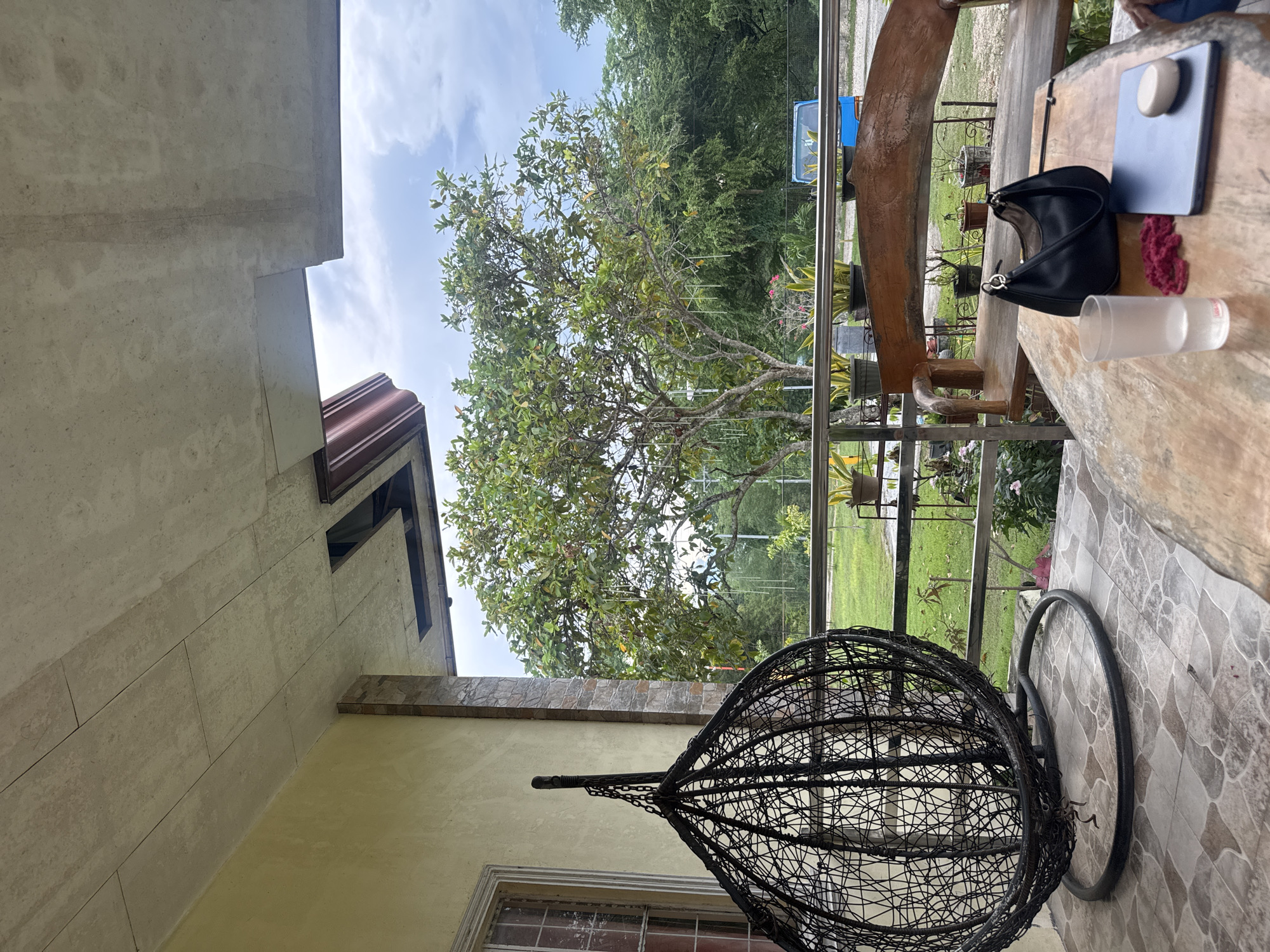 This is an outdoor photograph taken on the porch of a house in Samal, Philippines. The scene is viewed from a low angle, looking out towards a lush green landscape. In the foreground, a large, black, woven hanging chair with a metal stand occupies the left side of the frame. To the right, a rustic wooden table holds a glass of water, a black handbag, a laptop, and a small white object. Behind the table, a wooden bench with carved armrests faces the outdoors. A metal railing separates the porch from the yard, where several potted plants are arranged. Beyond the railing, a large tree with vibrant green foliage stands prominently, its branches reaching towards a cloudy sky. In the distance, a bright blue vehicle is visible among other greenery. The house wall, painted a pale yellow, forms the left boundary of the porch, with a portion of a window visible. The ceiling of the porch is made of concrete panels, with visible gaps and unfinished sections. The flooring on the porch is tiled with a mosaic pattern. The overall atmosphere is one of a casual, relaxed outdoor setting.