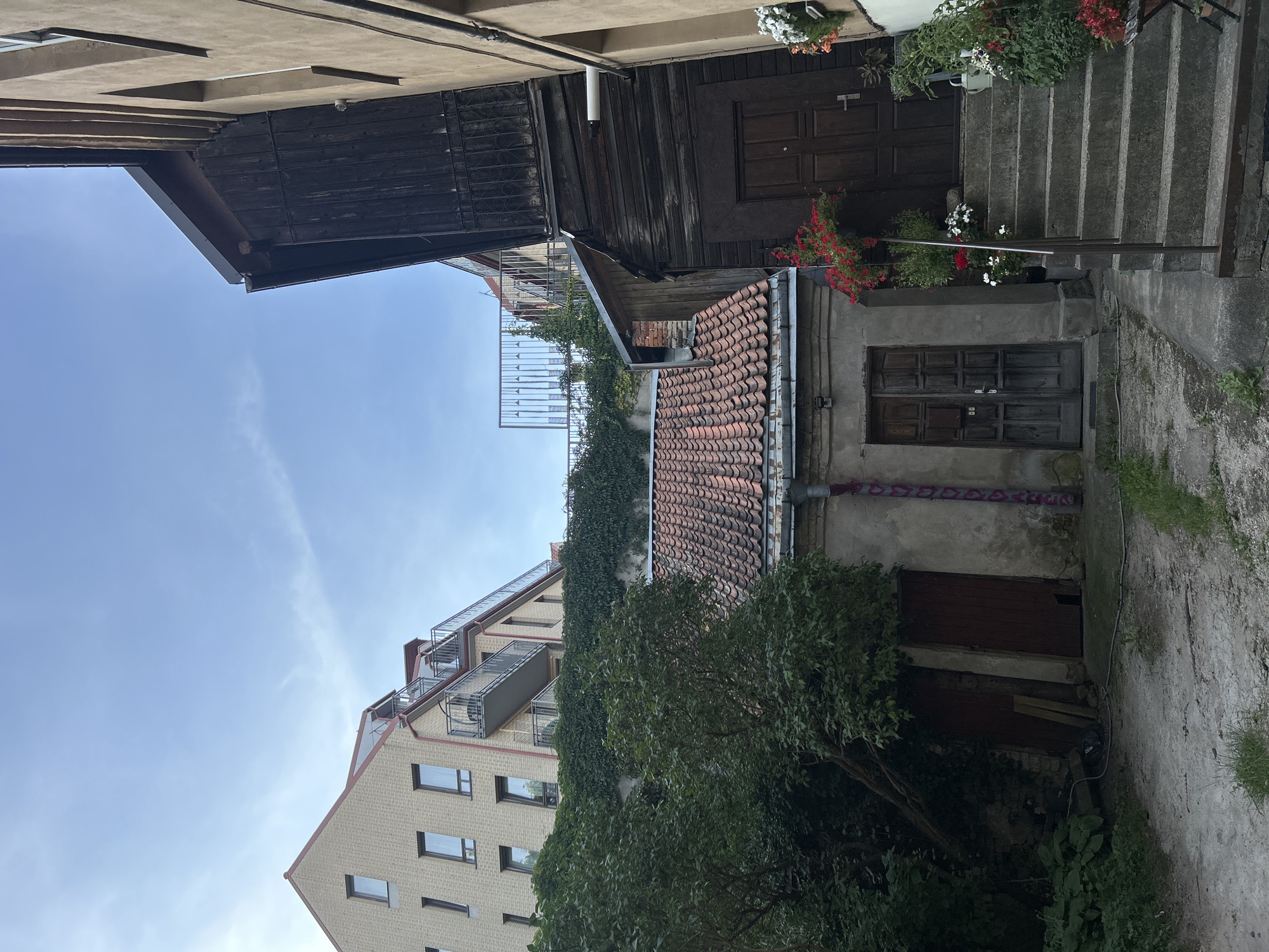 The image captures a courtyard area with multiple structures, suggesting an urban setting in Vilnius, Lithuania. On the left, a modern, light-colored brick building with several windows and balconies is visible, partially obscured by lush green foliage. In the center, a lower, older building with a red-tiled roof and weathered grey walls features two wooden doors. To the right, a dark wooden building with a steep roofline is prominent, with a wooden door at the bottom of a concrete staircase. Red and white flowers in pots are placed along the stairs and the entrance of the wooden structure. The ground is a mix of dirt, gravel, and some patches of grass. The sky above is a pale blue with wispy clouds. The scene appears to be a residential or mixed-use area, possibly in a historic part of the city, showing a contrast between older and newer architectural styles. There are no people or animals visible in the image.