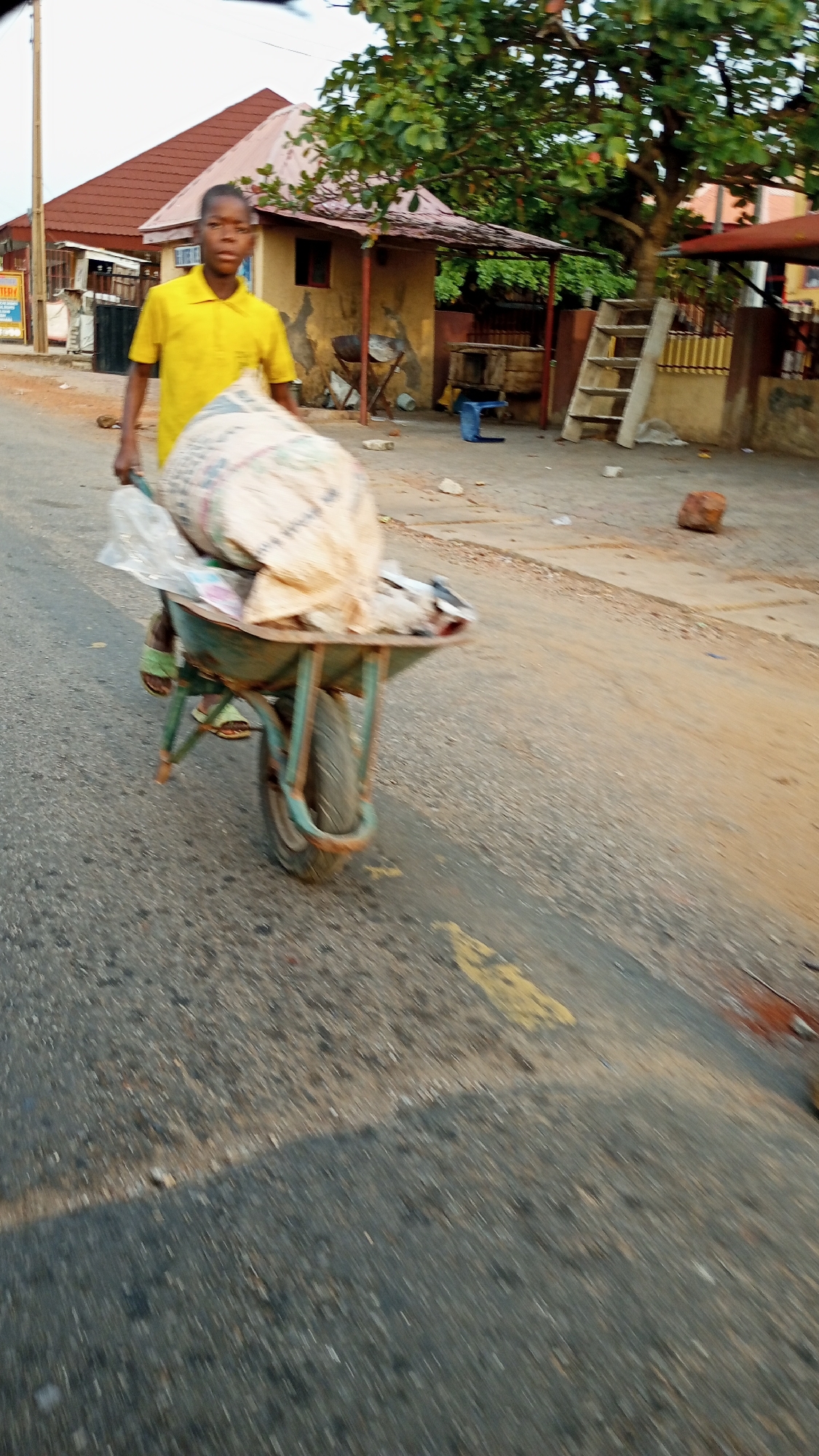 A young person in a yellow shirt is pushing a wheelbarrow loaded with a large bag down a paved road. The environment includes buildings, trees, and other roadside objects. The location appears to be a residential area in an unidentified city, country unknown.