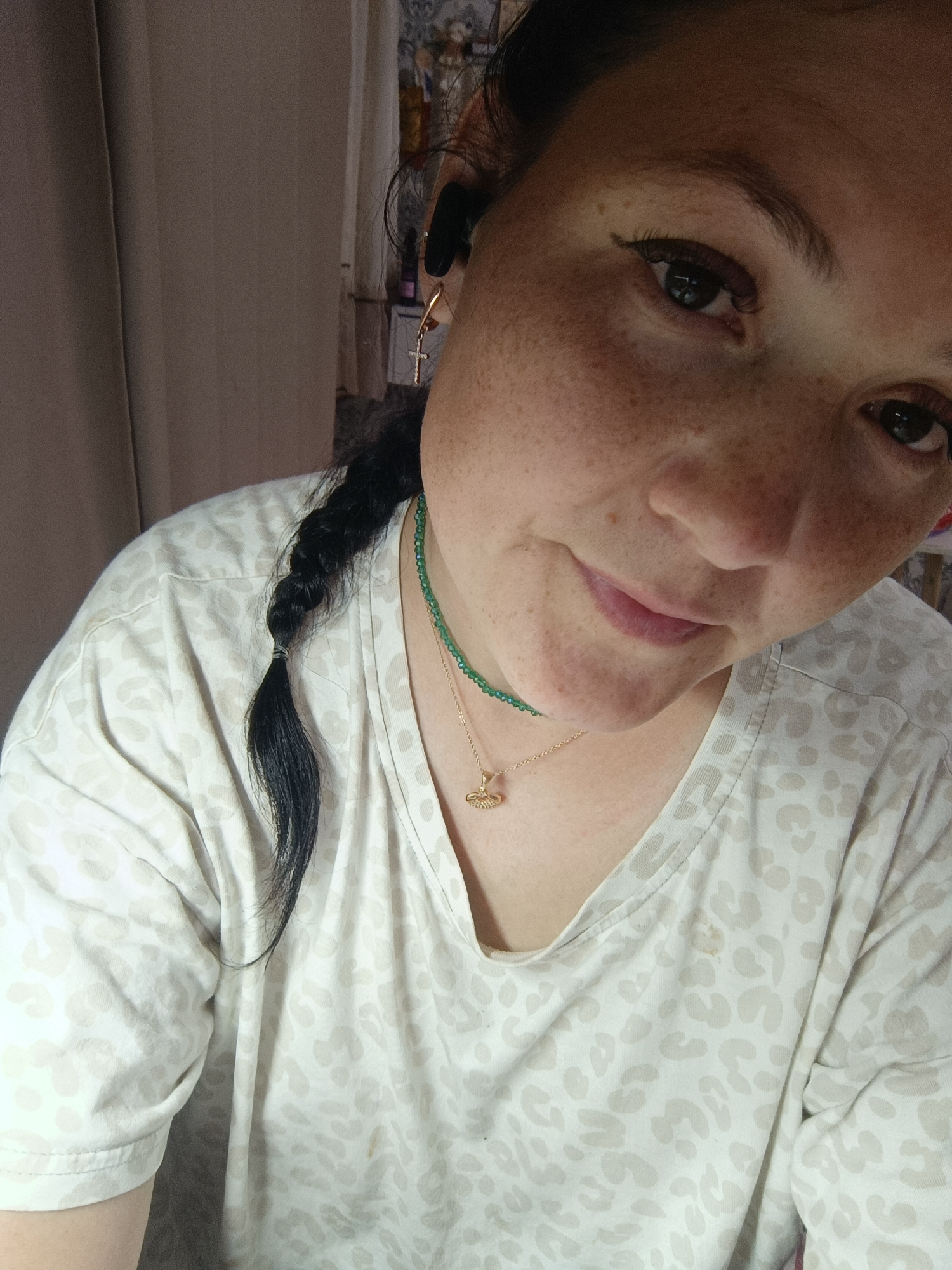 A close-up, overhead shot shows a woman with dark hair in a braid, wearing a light-colored t-shirt with a leopard print. She has freckles on her face and wears subtle makeup, including eyeliner and mascara. A green beaded necklace and a gold chain with a pendant are visible around her neck. She is also wearing black earbuds. The background is softly lit and features a neutral-colored curtain. The image does not provide any specific information about the city of Chaikovsky, Russia.