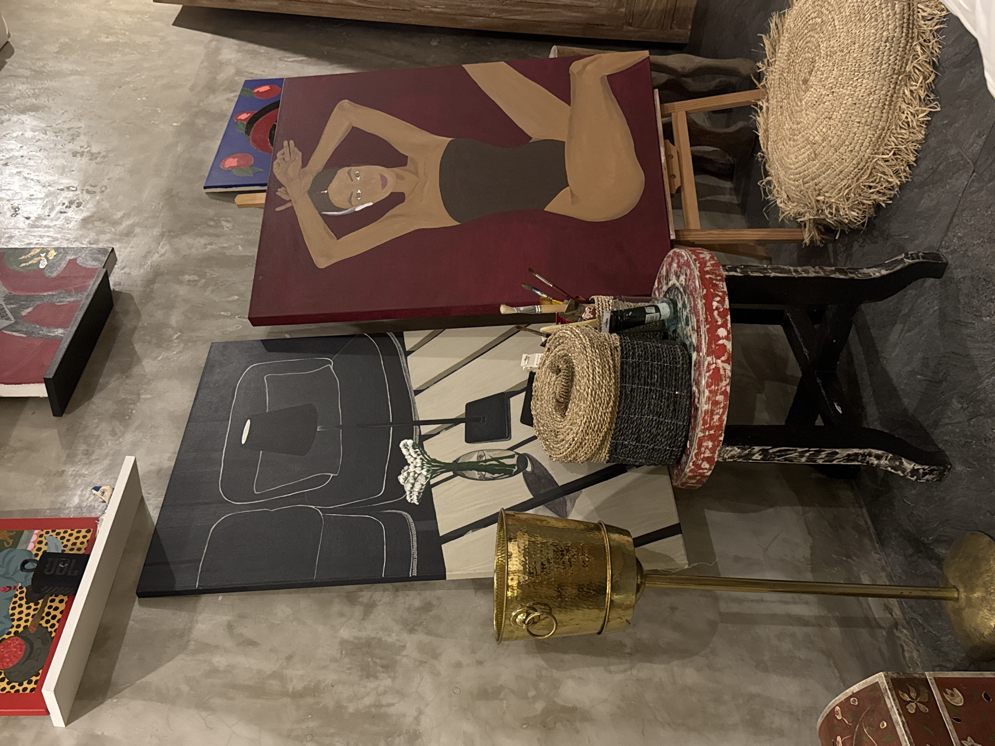 The image captures an indoor scene, likely an art studio, gallery, or creative workspace, with several artworks and crafting materials arranged on a concrete floor. The setting appears informal, with items displayed and stored.
The prominent subject is a large painting on an easel, depicting a reclining woman in a dark swimsuit against a deep maroon background. She has light brown skin, dark hair, and distinctive white-rimmed eyes. Her arms are raised above her head, and her expression is serene. Adjacent to this artwork, a collection of paintbrushes and tubes of paint suggests ongoing artistic activity.
Below the maroon painting, another large artwork features a stylized, abstract design in black and white, including outlines of figures and a vase holding white flowers. To the left, several smaller paintings are visible; one showcases a colorful, patterned design on a red background, with "JBL" faintly visible on a black object within the composition. Another small blue painting with red circular motifs is stacked behind the maroon one.
Among the objects on the floor are a small, dark-colored stool with a red and white painted top, a large circular woven mat with a fringed edge, and rolls of natural fiber material, possibly straw or rope. A brass-colored bucket or container, potentially part of a floor lamp given its elongated base, stands upright. The overall ambiance is artistic and somewhat rustic, characteristic of a creative environment. The lighting is artificial, casting subtle shadows on the floor.