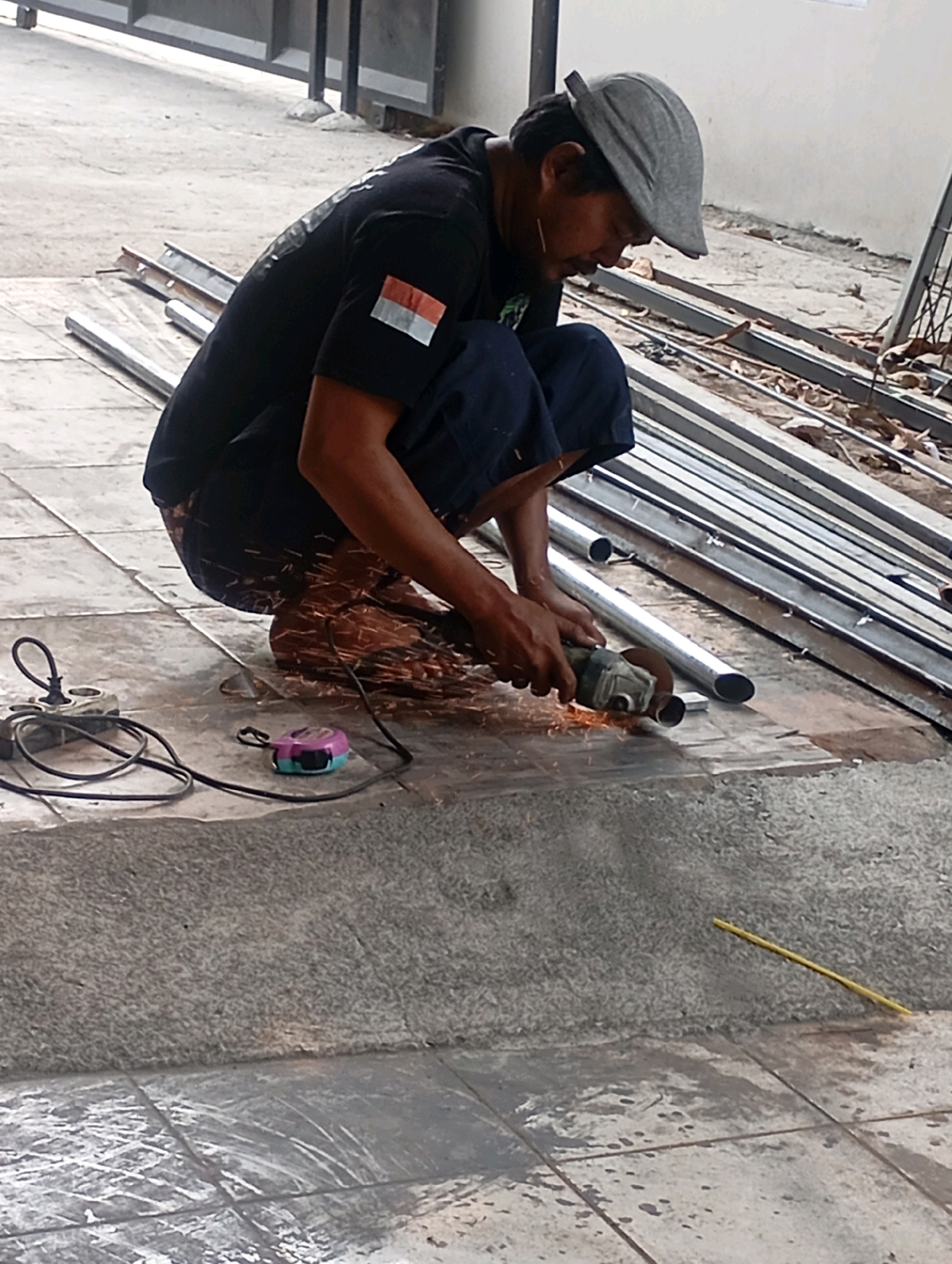 In the city of Babelan, Indonesia, a man wearing a grey flat cap and a black t-shirt with an Indonesian flag patch on the sleeve is seen crouching on a tiled surface. He is actively using an angle grinder to cut a metal pipe, generating numerous sparks. Scattered around him are more metal pipes and construction materials, with fallen leaves visible on the ground. A power cord from the angle grinder is visible, as well as a tape measure with purple and teal accents, and another electrical tool with a cord. The scene appears to be an outdoor construction or workshop area.