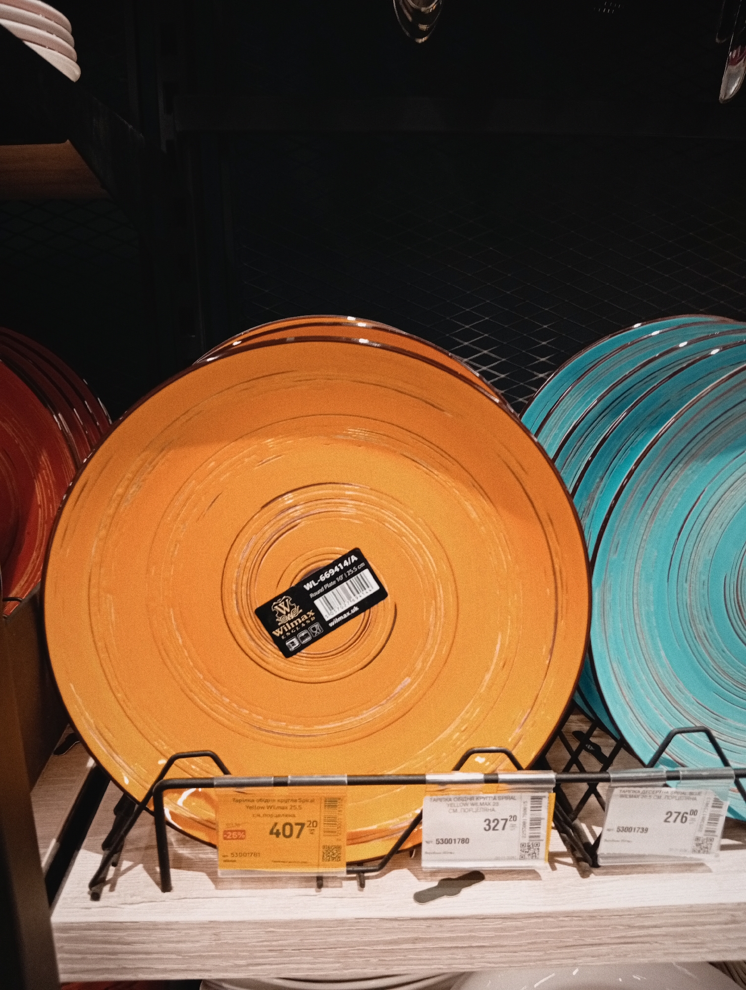 The image shows a display of colorful ceramic plates on a metal rack in a store. The plates are arranged in stacks, with a large orange plate in the center, a red plate to its left, and a blue plate to its right. Each plate has a price tag attached to the rack. The price tags indicate that the plates are manufactured by Wilmax. The plate closest to the camera, in the center, has a yellow price tag reading "407.20" with "-25%" printed next to it. The second plate has a price tag reading "327.20" and the last price tag reads "276.00". A label with "Wilmax England" is attached to the center of the plate closest to the camera. Based on the Cyrillic text and price tags, the location of the image is likely Poltava, Ukraine.