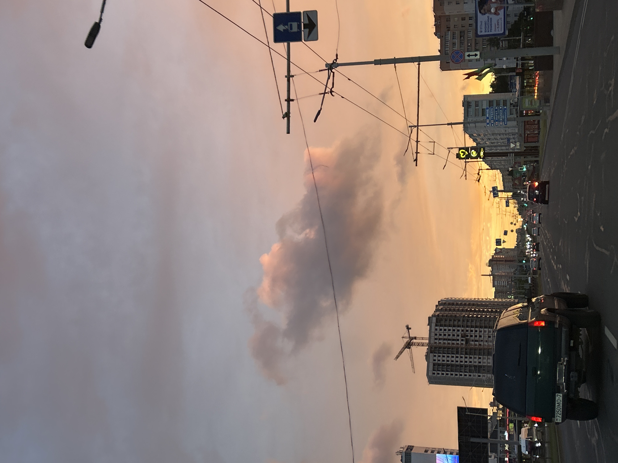 This image captures a street scene in Minsk, Belarus, during a sunset. The sky is filled with hues of orange, pink, and purple, with scattered clouds. A dark green SUV is in the foreground, driving down a multi-lane road. The road is wet, suggesting recent rain. On the right side of the road, there are streetlights, traffic signals, and various buildings. One building under construction, with a crane, is visible on the left. Traffic signs, including a directional sign with an arrow and a circular sign with a red border and blue symbol, are present. The overall atmosphere is urban and illuminated by the warm, fading sunlight.