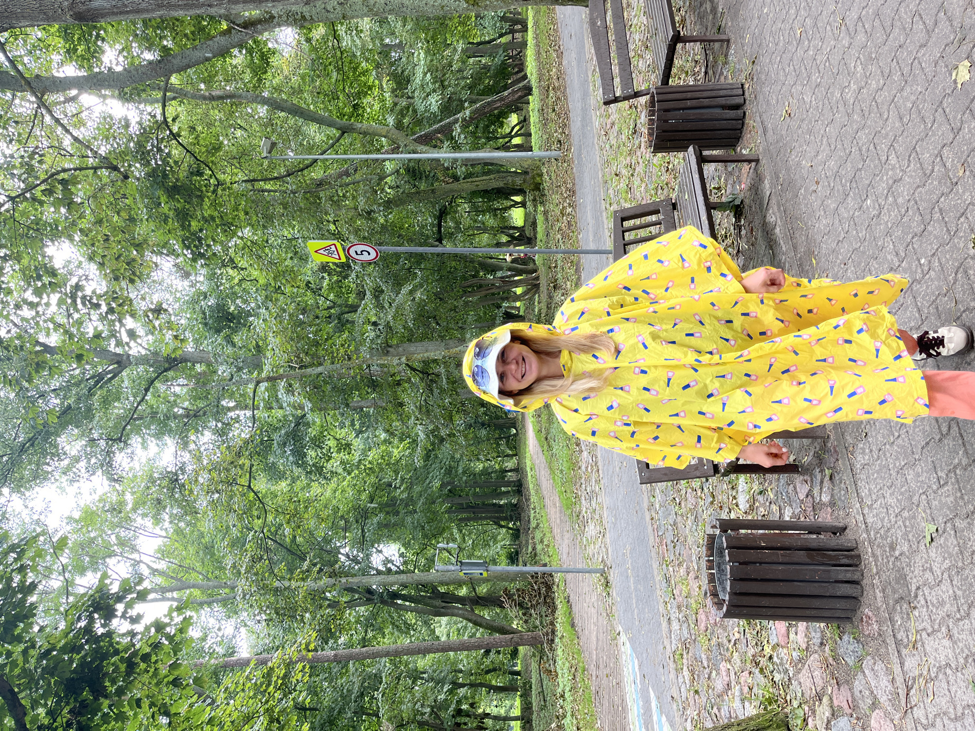 A woman in a yellow raincoat with a nail polish pattern stands in a park in Zelenogradsk, Russia. She is wearing a white bucket hat and sunglasses. She is smiling at the camera. In the background, there are trees, a paved path, benches, and trash cans. There are also two traffic signs, one indicating a pedestrian crossing and the other with the number 5. The ground is covered in cobblestones and fallen leaves.