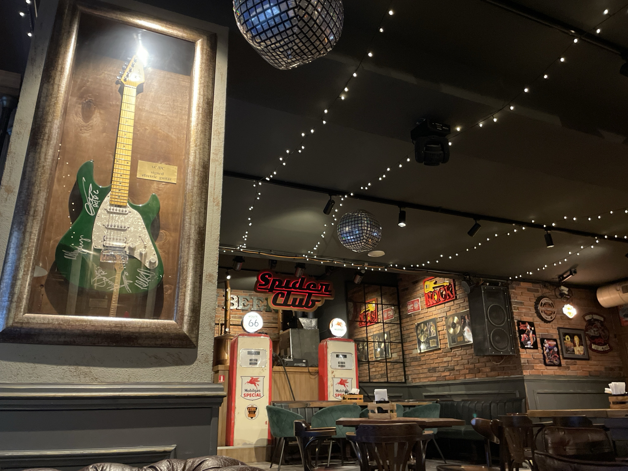 The image depicts the interior of a bar or restaurant, possibly in Veliko Tarnovo, Bulgaria. A framed, green electric guitar signed by AC/DC hangs on the wall. Disco balls and string lights illuminate the dark ceiling. The interior features a brick wall decorated with music memorabilia, including images of musicians, gold records, and a Route 66 sign. A large speaker is also attached to the wall. Two vintage Mobilgas Special gas pumps stand near the bar, adding to the retro aesthetic. Tables and chairs are arranged for customers. The overall atmosphere is reminiscent of a rock-themed establishment.