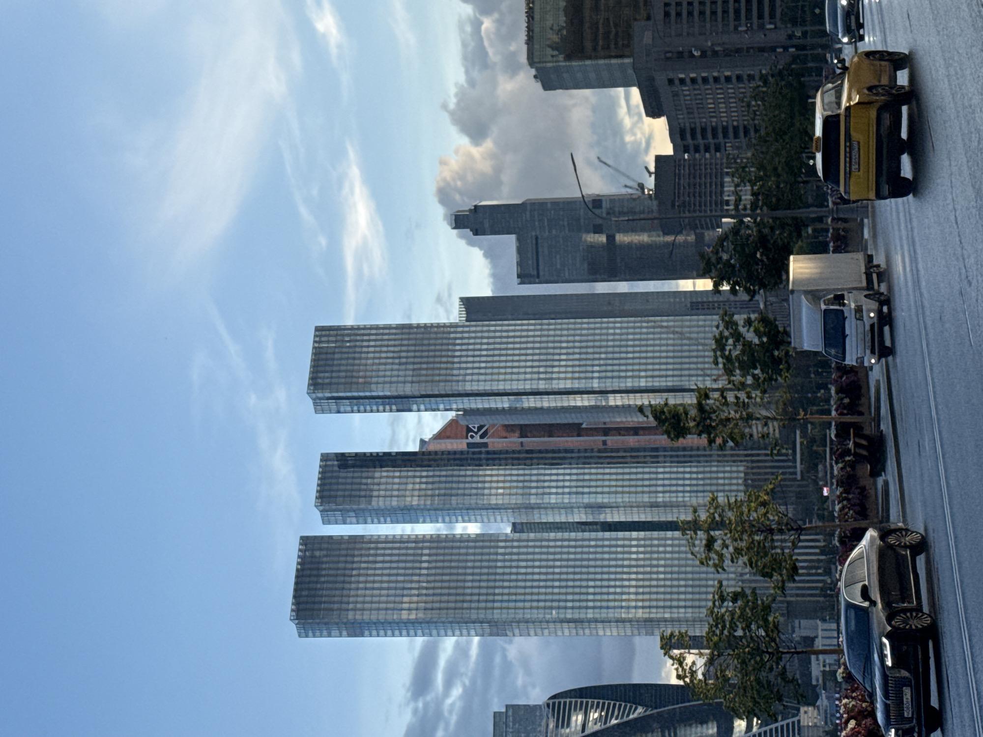 The image depicts a street view of the Moscow City business district in Russia. In the foreground, a black sedan is seen driving on a wet road, its headlights illuminated. To its right, a yellow SUV, possibly a taxi, is also traveling on the road. Behind them, a white truck follows. The background is dominated by several modern skyscrapers, characterized by their glass facades. The sky above is a mix of blue with scattered clouds, and some trees are visible along the side of the road, suggesting a park or landscaped area. The overall scene portrays an urban environment with active traffic and contemporary architecture.