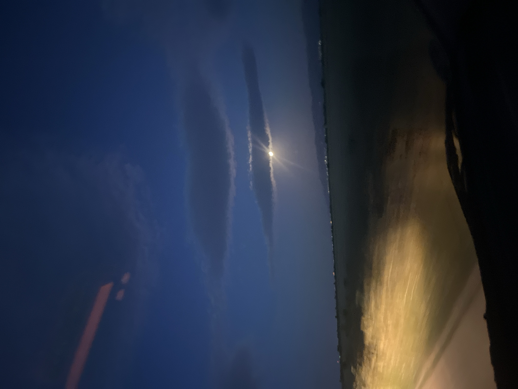 A nighttime view from inside a vehicle shows a scene in an unknown location. The sky is a dark blue with elongated, cloud-like formations. A bright light source, possibly the moon, is visible through the clouds. The right side of the frame features the side of the vehicle's interior, reflecting a yellowish light, likely from the vehicle's headlights, onto a surface. Along the horizon line, small points of light suggest either a cityscape or reflections on water. The image orientation is sideways.