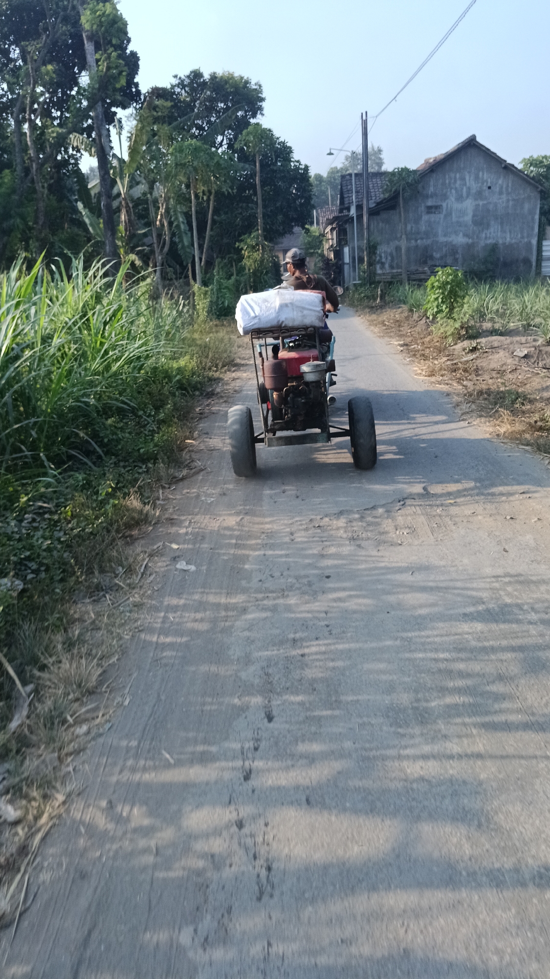 A person wearing a cap is driving a red and black motorized agricultural vehicle with two large rear wheels and two smaller front wheels down a paved road in the village of Slumbung, Indonesia. The vehicle is carrying a large white bundle on a rack behind the driver. Lush green vegetation lines the left side of the road, while houses with gray concrete walls are visible on the right. The road is partially shaded by trees, casting shadows on the surface. The sky is clear and pale blue.