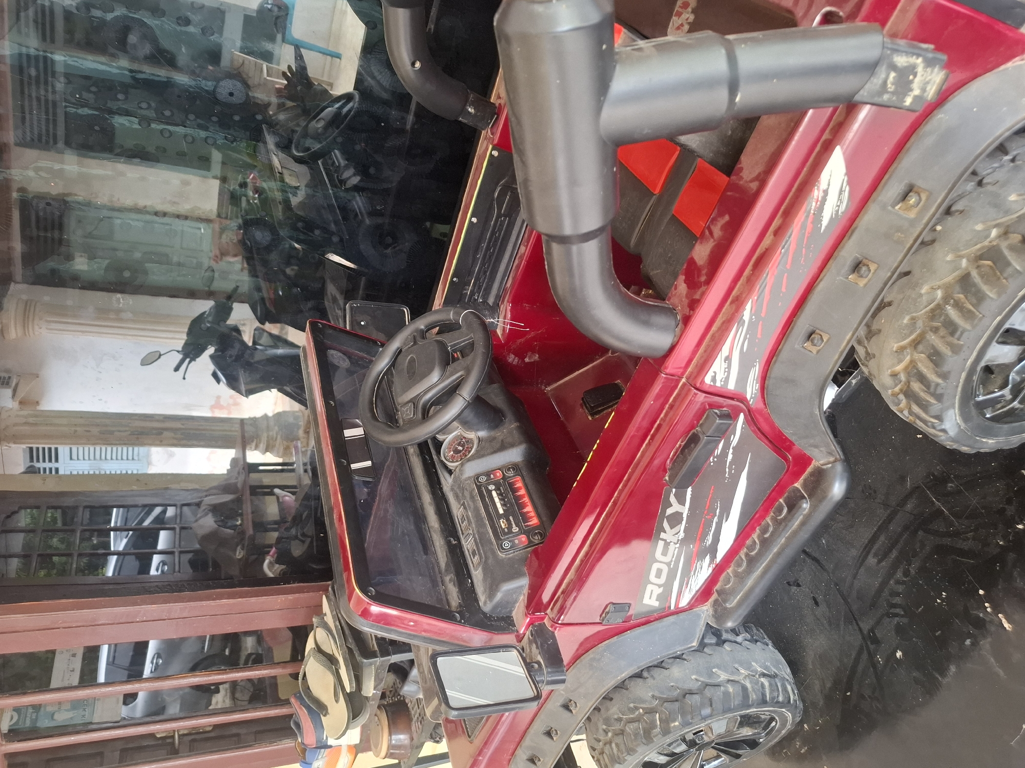 The image shows a red, toy Jeep with the word "ROCKY" on its side, parked on a black, reflective surface. The Jeep has large, rugged tires, a steering wheel, and a dashboard with buttons and a small screen. In the background, there's a building with glass doors and windows, through which motorcycles and a silver car are visible. The setting appears to be outdoors, possibly a driveway or porch, in a location that suggests a tropical climate due to the foliage and architecture. The specific location is identified as Deah Pangwa, Indonesia.