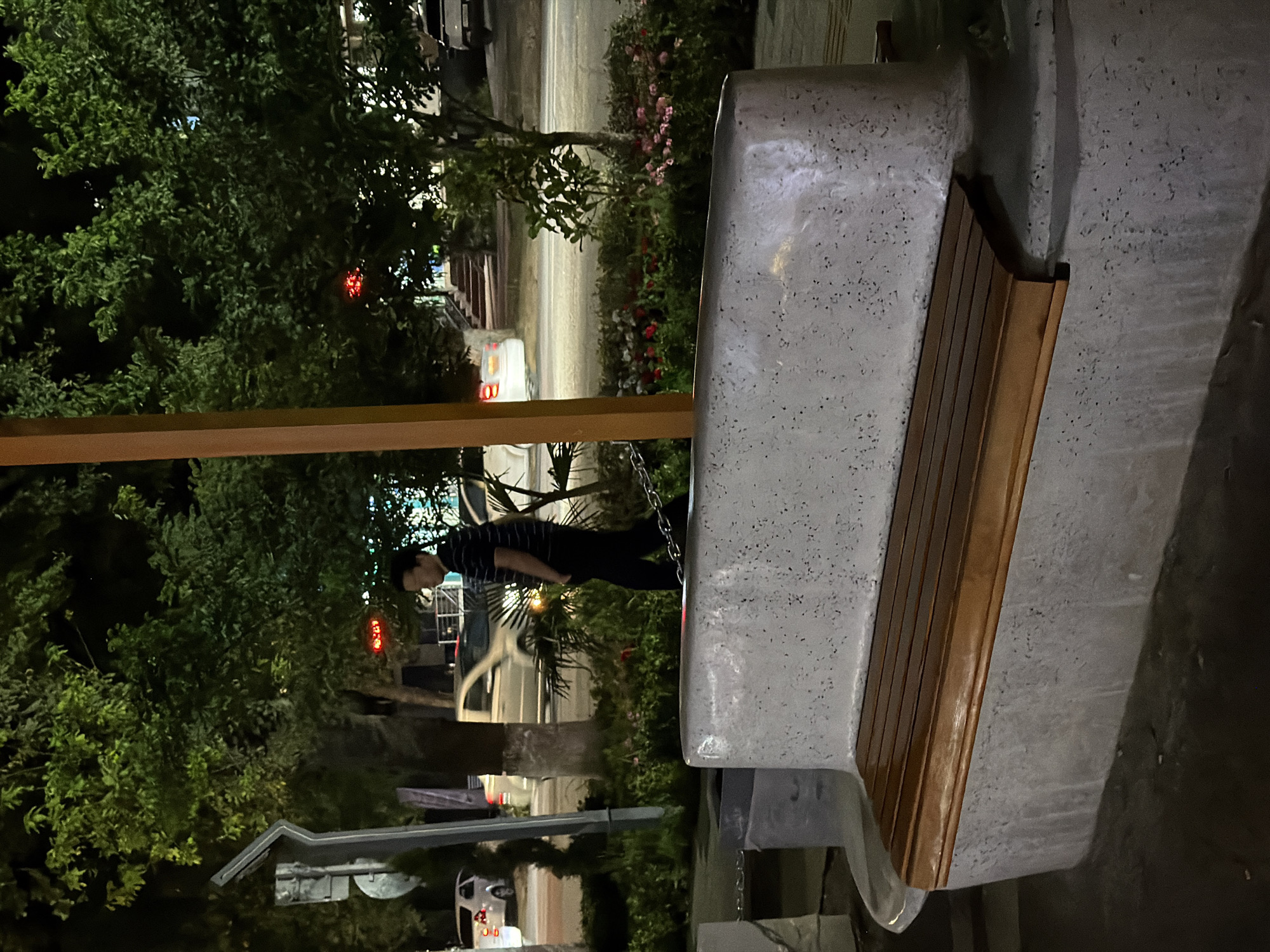A person stands beside a concrete bench with wooden slats, outdoors at night. The person, wearing a striped shirt and dark pants, is leaning forward and looking down. The bench is placed on a paved area, with a chain extending from its base. Behind the bench, a tree with dense foliage is visible, with some red lights or flowers in the background. Further back, a street scene is dimly lit, with vehicles and buildings suggested. The overall impression is one of a public space at night, possibly a park or a street lined with trees. The lighting is artificial, casting shadows and creating a slightly moody atmosphere. The location context of Chirchik, Uzbekistan, is not explicitly evident from the visual cues alone, but it provides a geographical anchor for the scene. There is no visible text within the image.