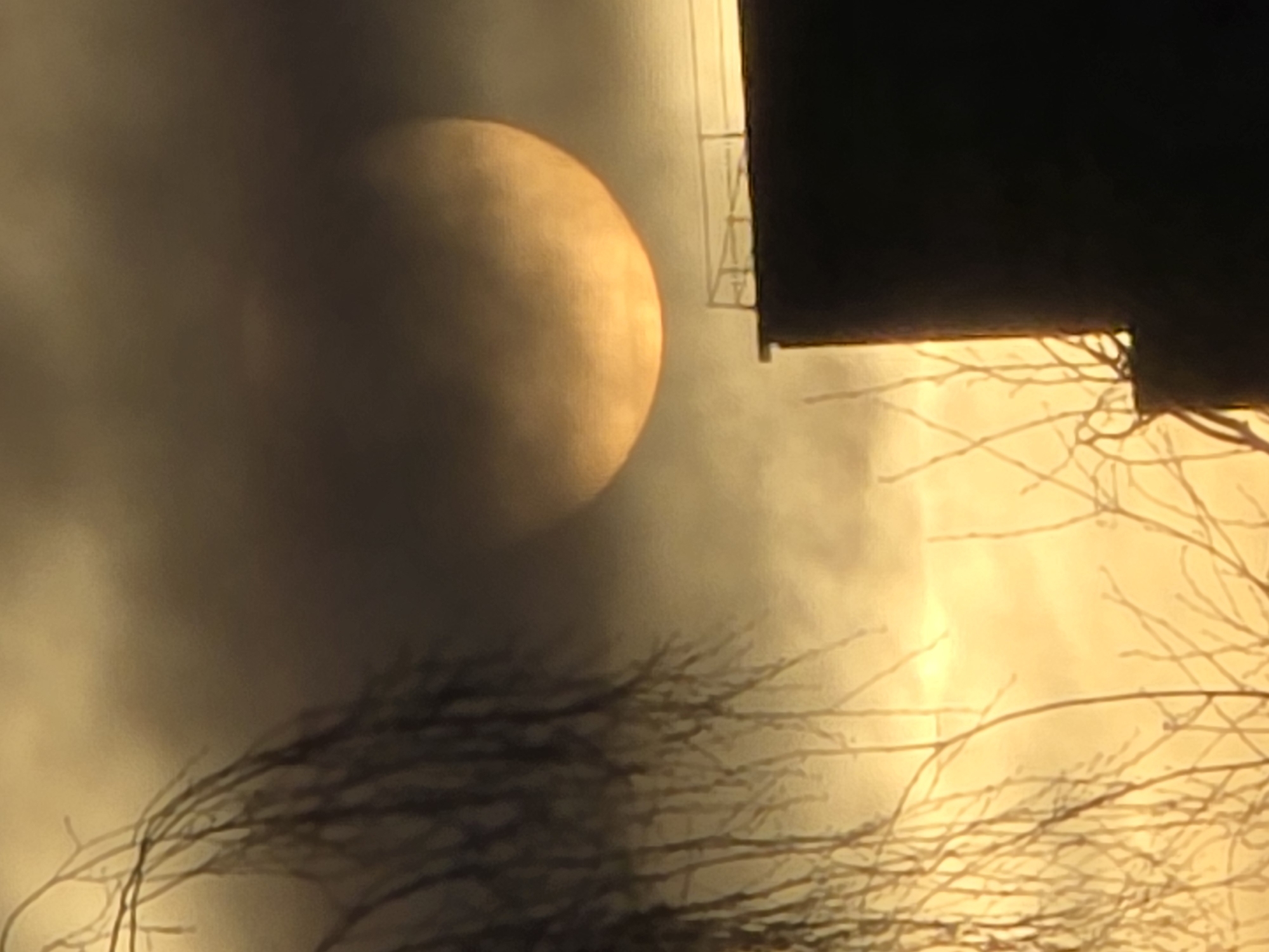 The image shows a large, blurred orange disc, possibly the sun, obscured by clouds or smoke. A dark, silhouetted structure, possibly a building, appears in the upper right, with some architectural details visible. Bare branches of trees are visible in the lower portion of the image. The scene appears to be a twilight or sunrise, with a warm, golden light filtering through the atmosphere. The location is Unknown, Unknown. No people are visible.