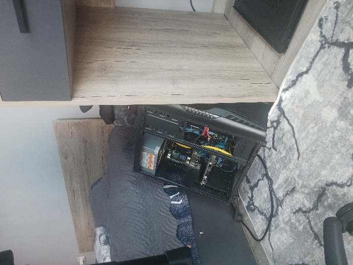 The scene depicts an interior room in Velichkovo, Bulgaria. In the center, a black computer tower, with its left side panel removed, rests on a light grey rug featuring dark grey irregular patterns. The internal components, including circuit boards, a visible fan, and electrical wiring in yellow, blue, and red, are exposed. A white warning label is affixed inside the case. To the left, a bed is visible with a grey quilted comforter and a blue and white patterned sheet, positioned beneath a light wood-grain headboard against a light grey wall. To the right, a light wood-grain desk or shelf unit, featuring a dark grey drawer front, partially obscures the computer tower. A black cable lies across the rug in the foreground. A black rectangular object, possibly a laptop or device stand, is situated on the floor beneath the desk unit. No individuals are present.